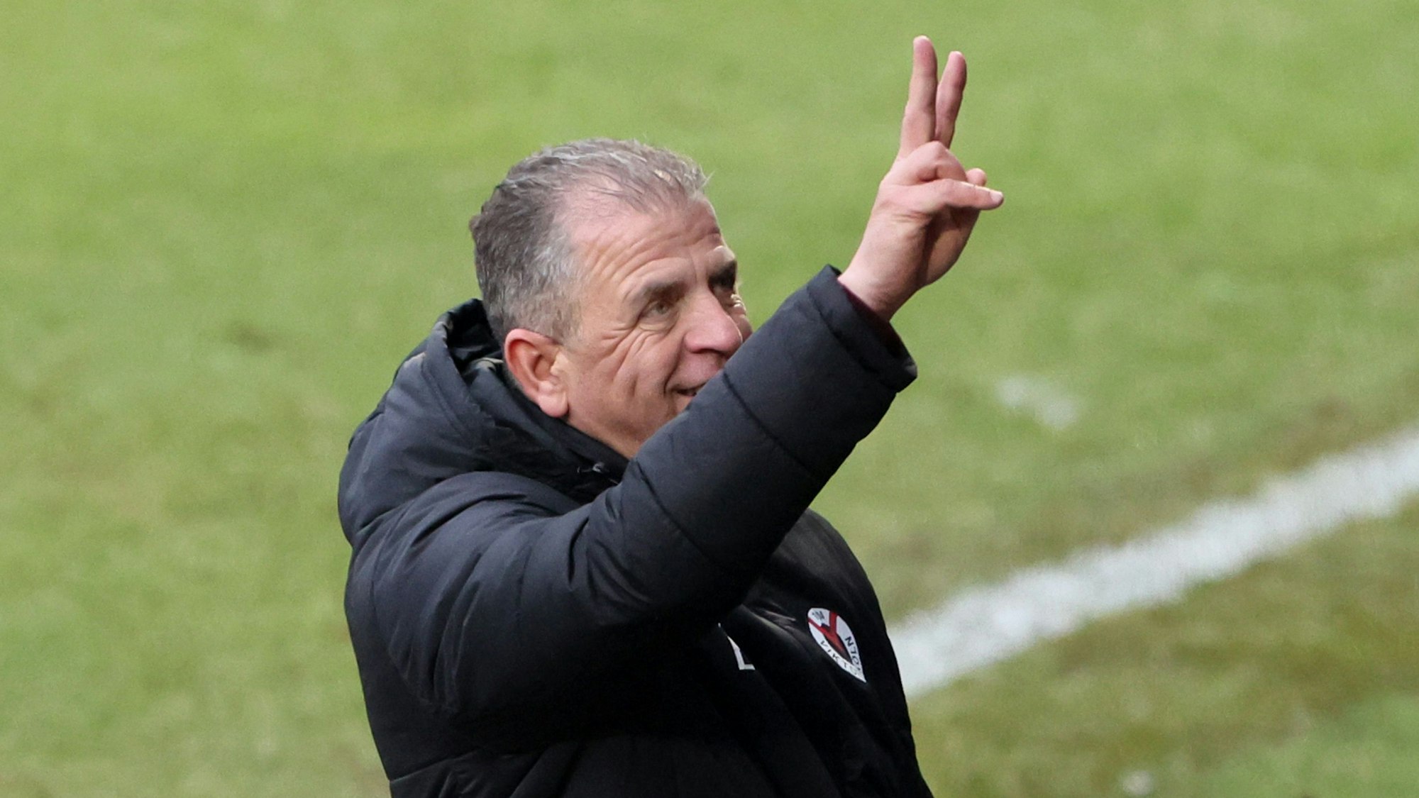 Ein Fußball-Manager steht am Spielfeldrand und grüßt jemanden auf der Tribüne.