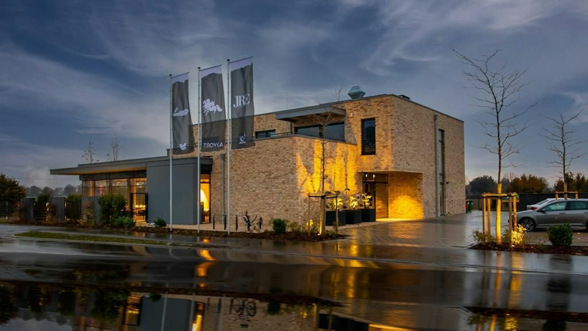 quadratisches Gebäude, das im Halbdunkeln beleuchtet wird, rechts ein Parkplatz