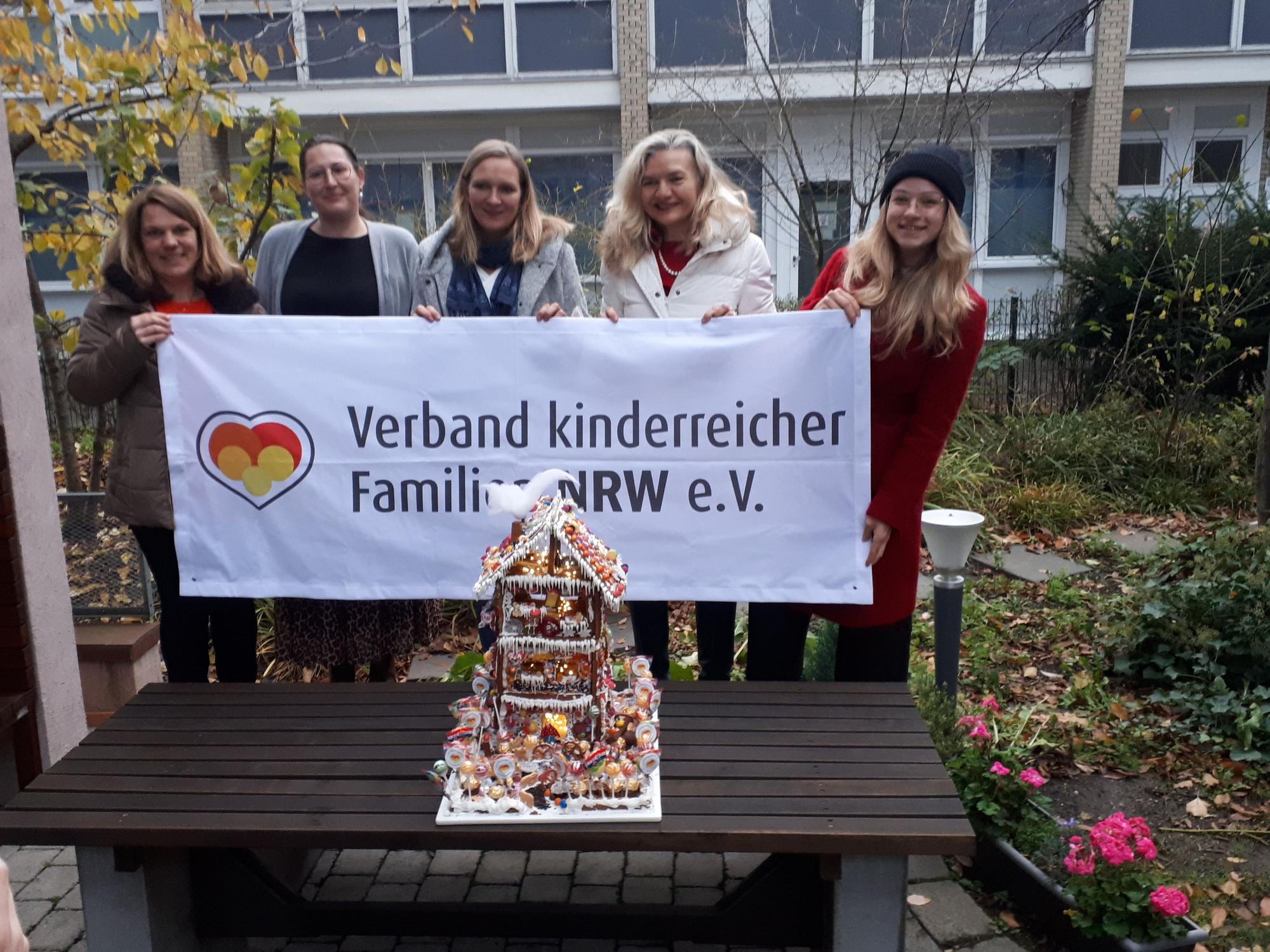 Fünf Frauen halten ein Transparent hoch mit dem Schriftzug Verband kinderreicher Familien NRW e.V. Auf einer Bank davor steht ein liebevoll verziertes vier Etagen hohes Lebkuchenhaus.