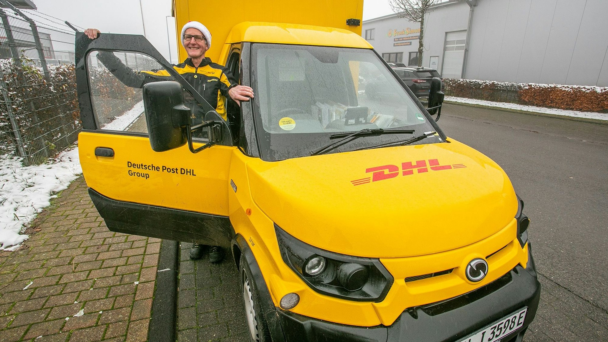 Ein Mann mit Mütze und einem Deutsche Post-Outfit steht in der Türe des gelben DHL-Transporters. Durch die Scheibe sieht man das Lenkrad auf der rechten Seite