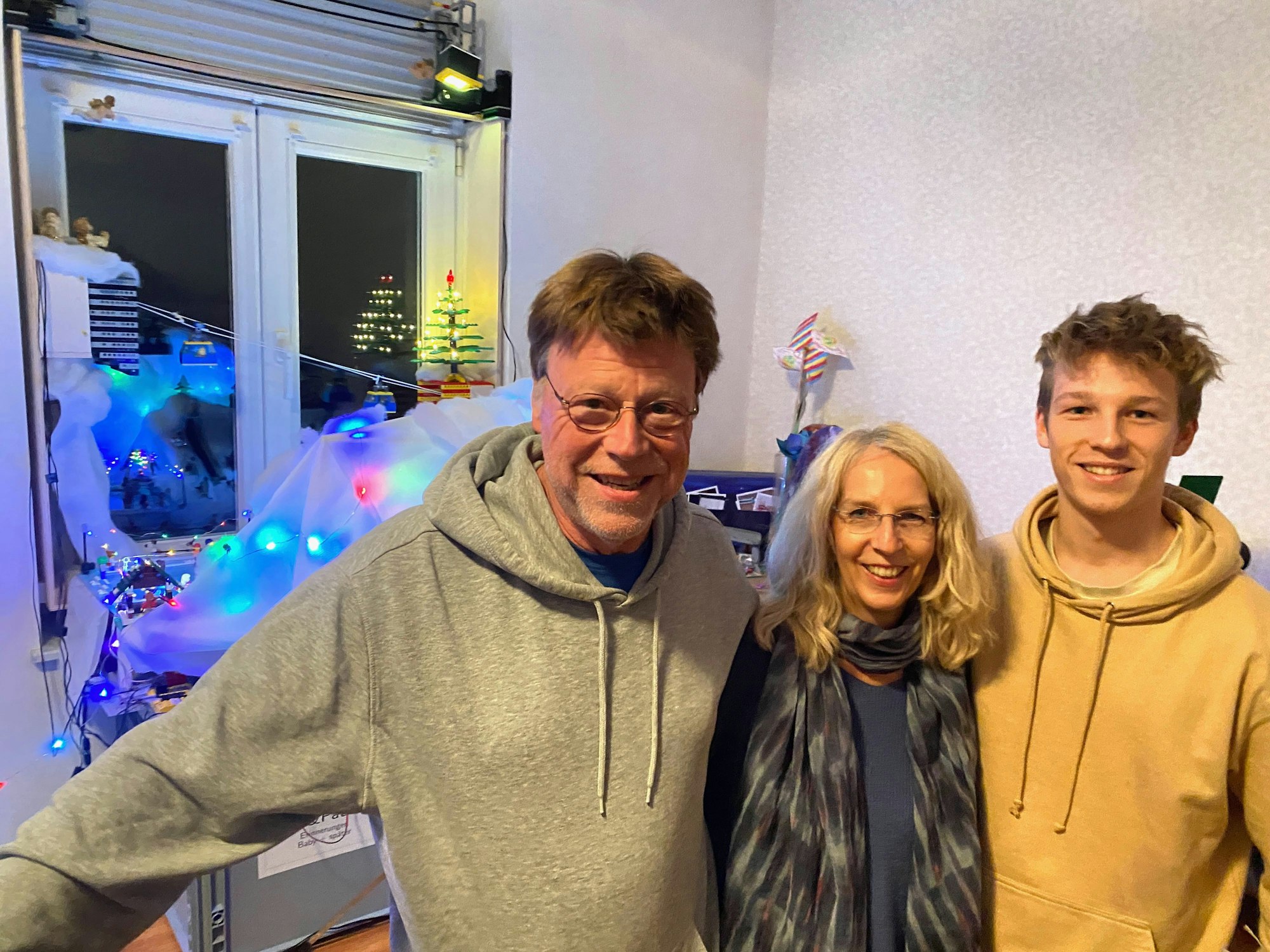 Ein Mann und eine Frau und ein jüngerer Mann, Eltern und Sohn, stehen nebeneinander, im Hintergrund sind mit Lichterketten beleuchtete Lego-Aufbauten zu sehen.