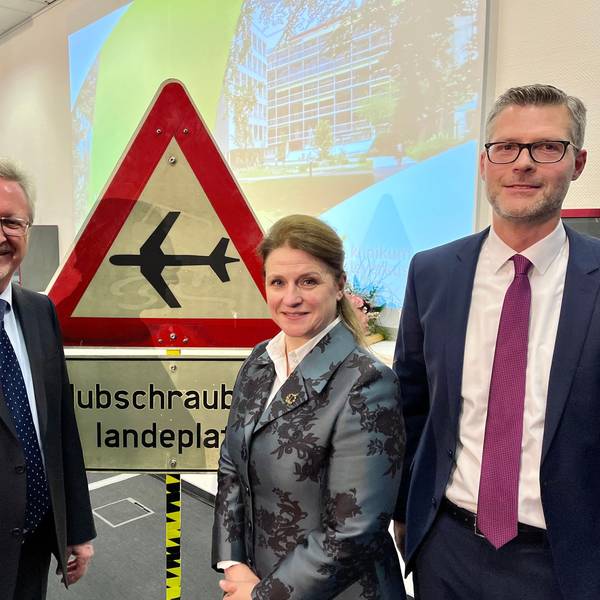 Hans-Peter Zimmermann mit seinen Nachfolgern Anja Mitrenga-Theusinger und André Schumann vor einem Schild: Hubschrauber-Landeplatz