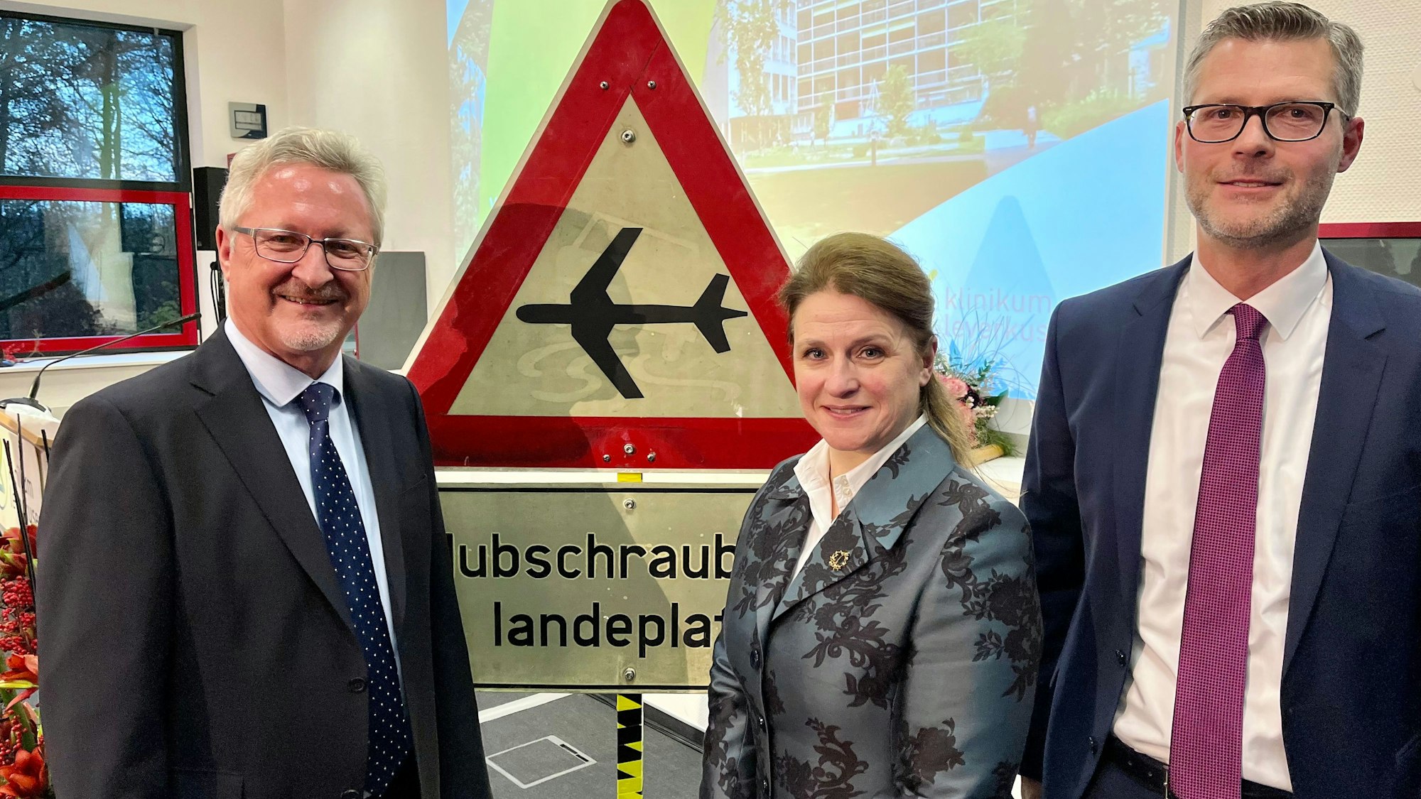 Hans-Peter Zimmermann mit seinen Nachfolgern Anja Mitrenga-Theusinger und André Schumann vor einem Schild: Hubschrauber-Landeplatz