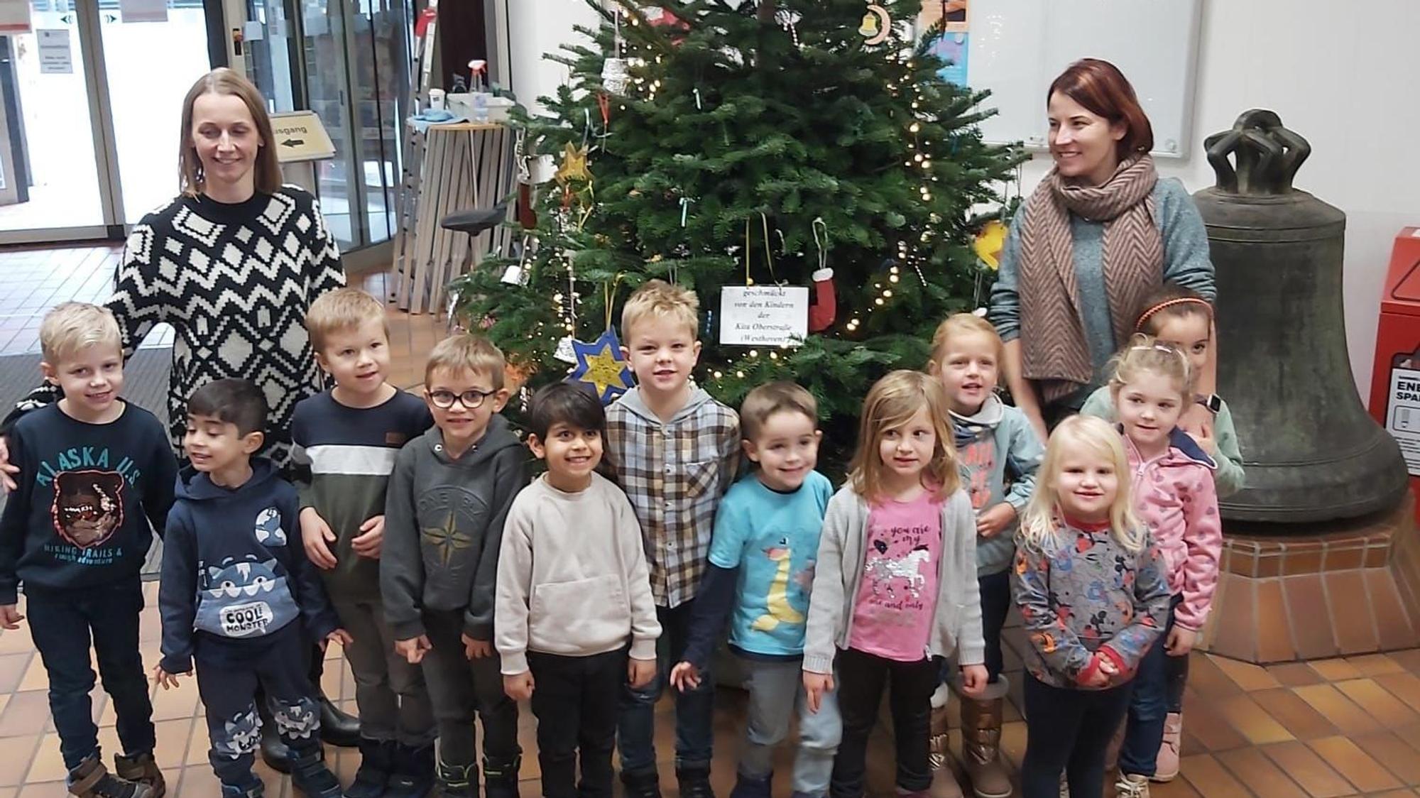 Kindergartenkinder und zwei Erzieherinnen stehen vor einem geschmückten Weihnachtsbaum im Foyer.