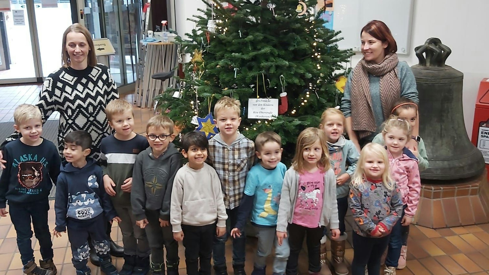 Kindergartenkinder und zwei Erzieherinnen stehen vor einem geschmückten Weihnachtsbaum im Foyer.