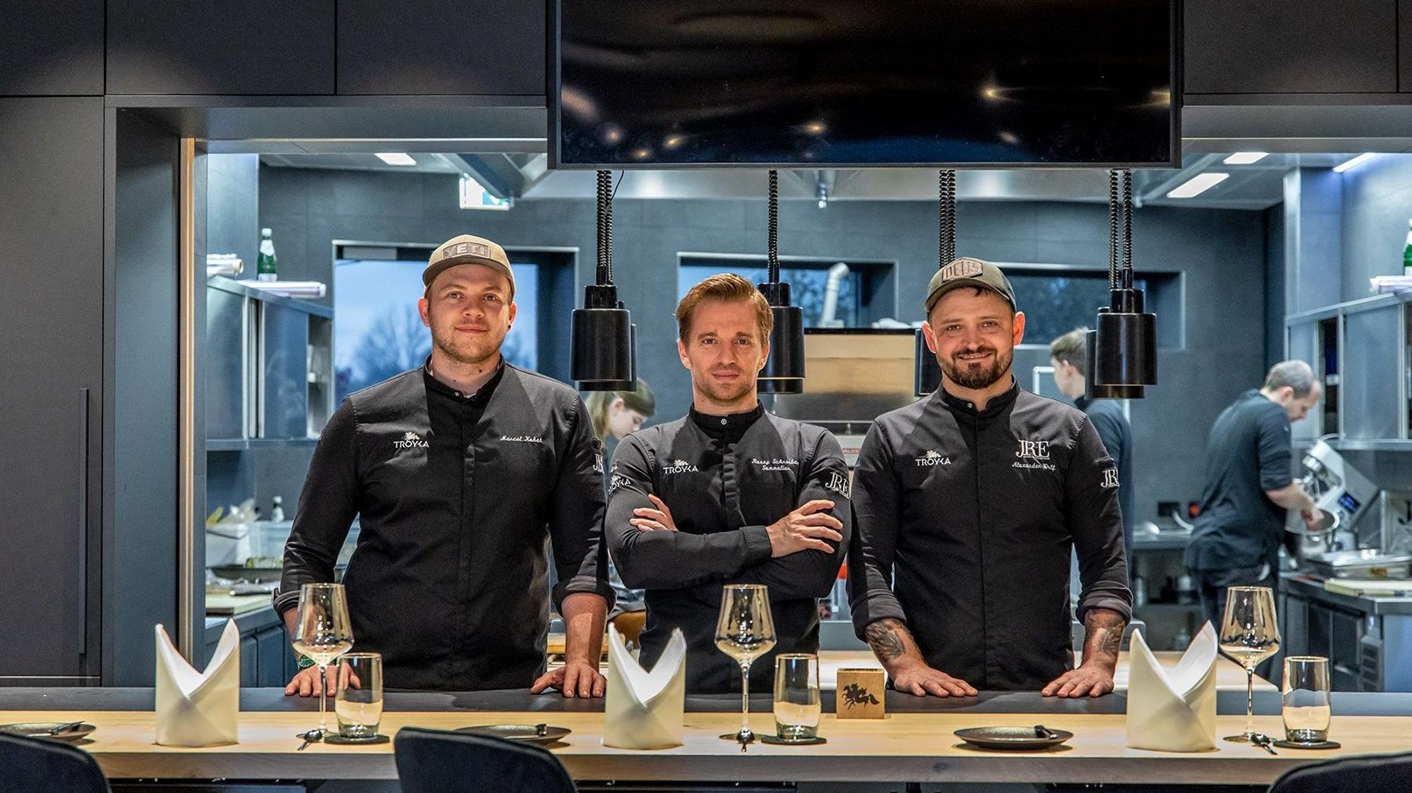 drei Männer in schwarzer Kochkleidung stehen vor einem gedecketen Tisch und blicken in die Kamera