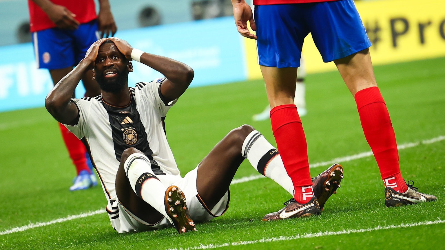 Der deutsche Nationalspieler Antonio Rüdiger sitzt auf dem Rasen während des WM-Spiels gegen Costa Rica. Er fasst sich an den Kopf. Außerdem sind die Beine von zwei costa-ricanischen Nationalspielern zu sehen.