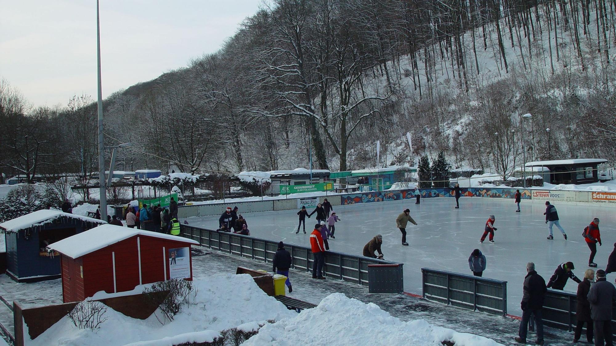 Eisbahn mit Schlittschuhläufern, verschneiter Wald im Hintergrund