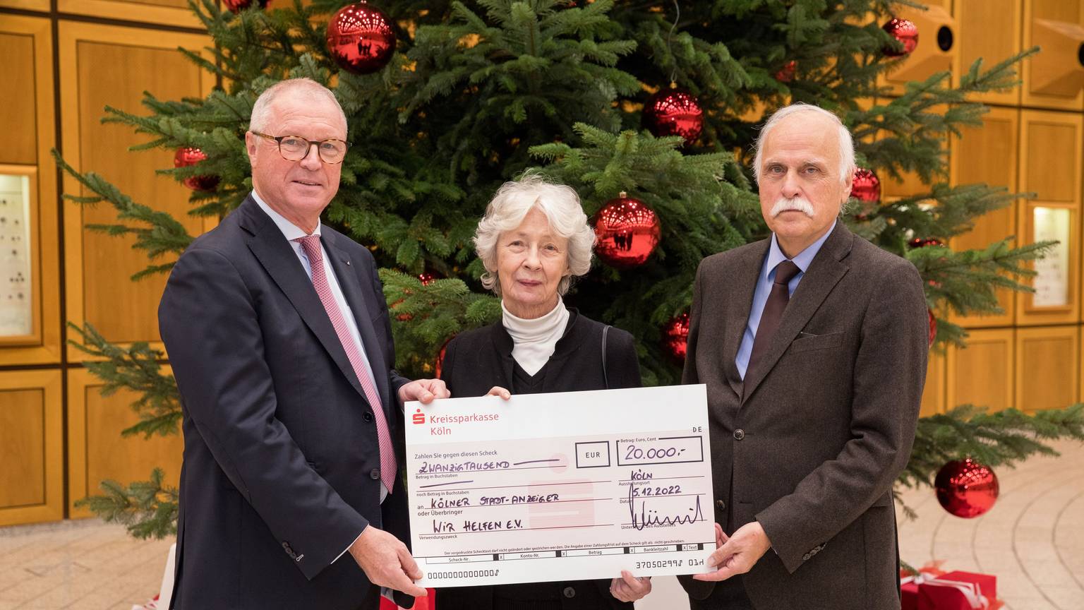 Der Vorstandsvorsitzende der Kreissparkasse Köln Alexander Wüerst überreicht vor einem großen Weihnachtsbaum einen symbolischen Scheck über 20.000 Euro an Hedwig Neven DuMont und Karl-Heinz Goßmann von „wir helfen“.