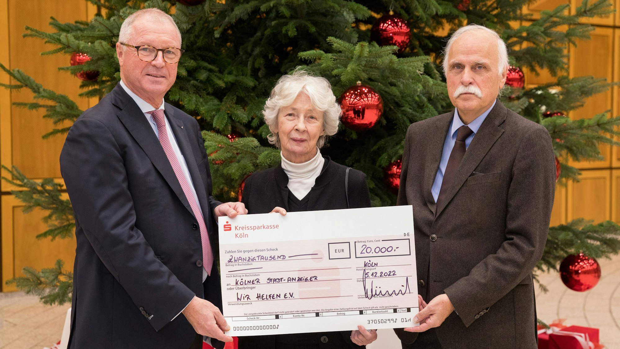 Der Vorstandsvorsitzende der Kreissparkasse Köln Alexander Wüerst überreicht vor einem großen Weihnachtsbaum einen symbolischen Scheck über 20.000 Euro an Hedwig Neven DuMont und Karl-Heinz Goßmann von „wir helfen“.