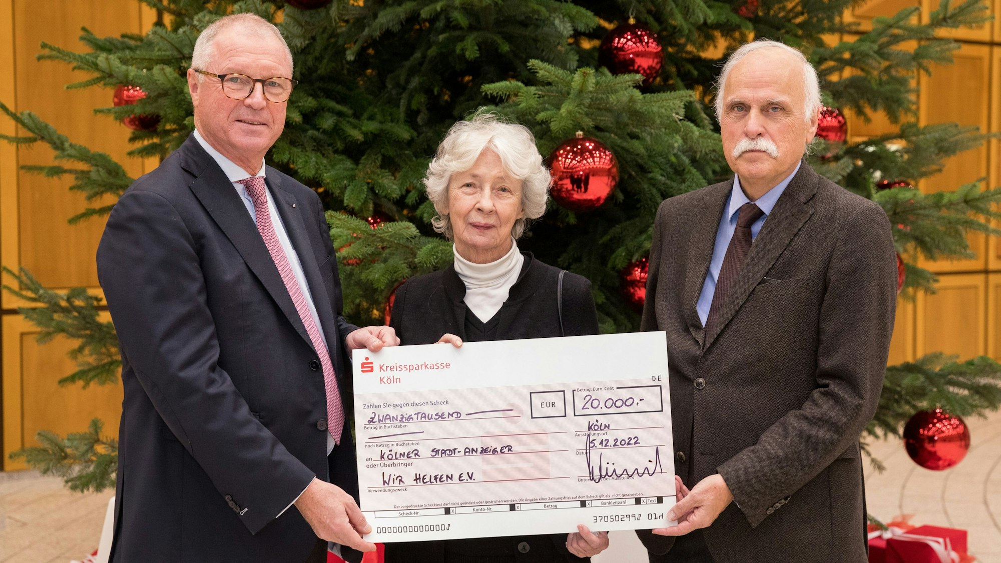 Der Vorstandsvorsitzende der Kreissparkasse Köln Alexander Wüerst überreicht vor einem großen Weihnachtsbaum einen symbolischen Scheck über 20.000 Euro an Hedwig Neven DuMont und Karl-Heinz Goßmann von „wir helfen“.