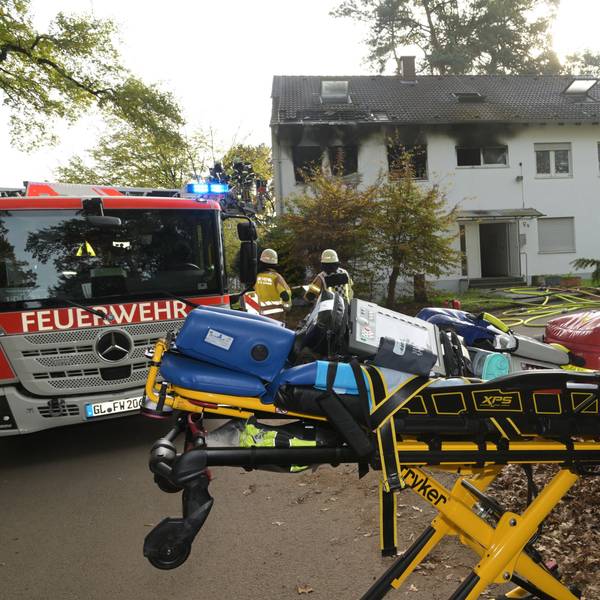 Rauch strmöt aus Fenster der Notunterkunft in Bergisch Gladbach, davor steht ein Feuerwehr und eine Bare mit Gegenständen darauf.