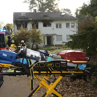 Rauch strmöt aus Fenster der Notunterkunft in Bergisch Gladbach, davor steht ein Feuerwehr und eine Bare mit Gegenständen darauf.
