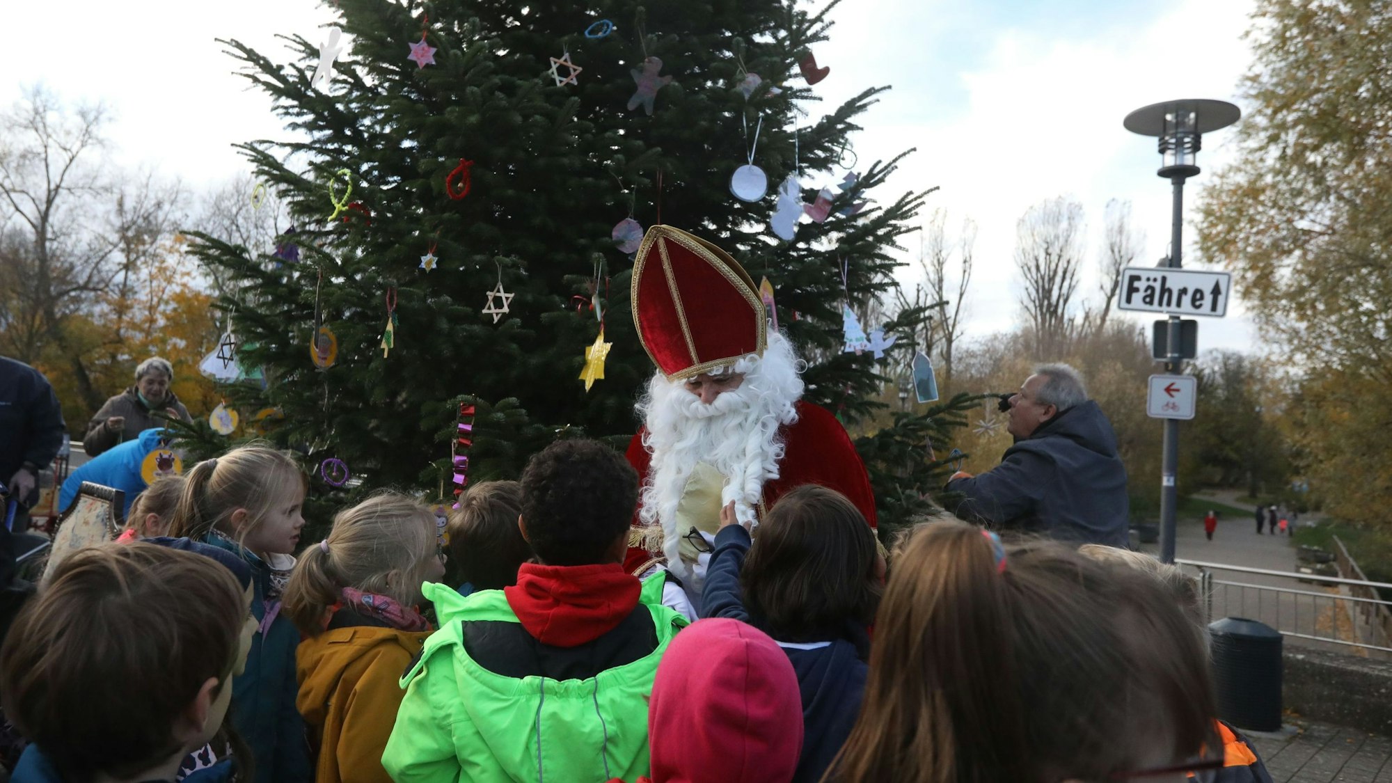 Kinder und eine als Nikolaus verkleidete Person stehen vor einem geschmückten Weihnachtsbaum.