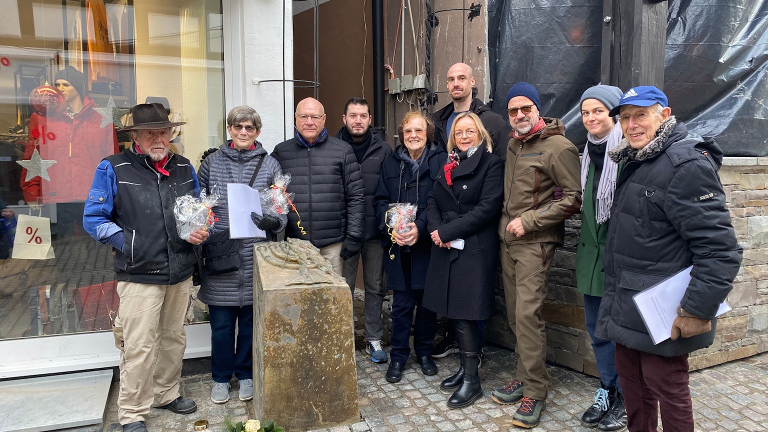 Künstler Gunter Demnig (l.) steht neben der Stele vor dem früheren Synagogenstandort in Bad Münstereifel. An ihrem Fuß sind weiße Rosen abgelegt. Unter den Teilnehmern der Aktion ist auch die Bad Münstereifeler Bürgermeisterin Sabine Preiser-Marian (4.v.r.).