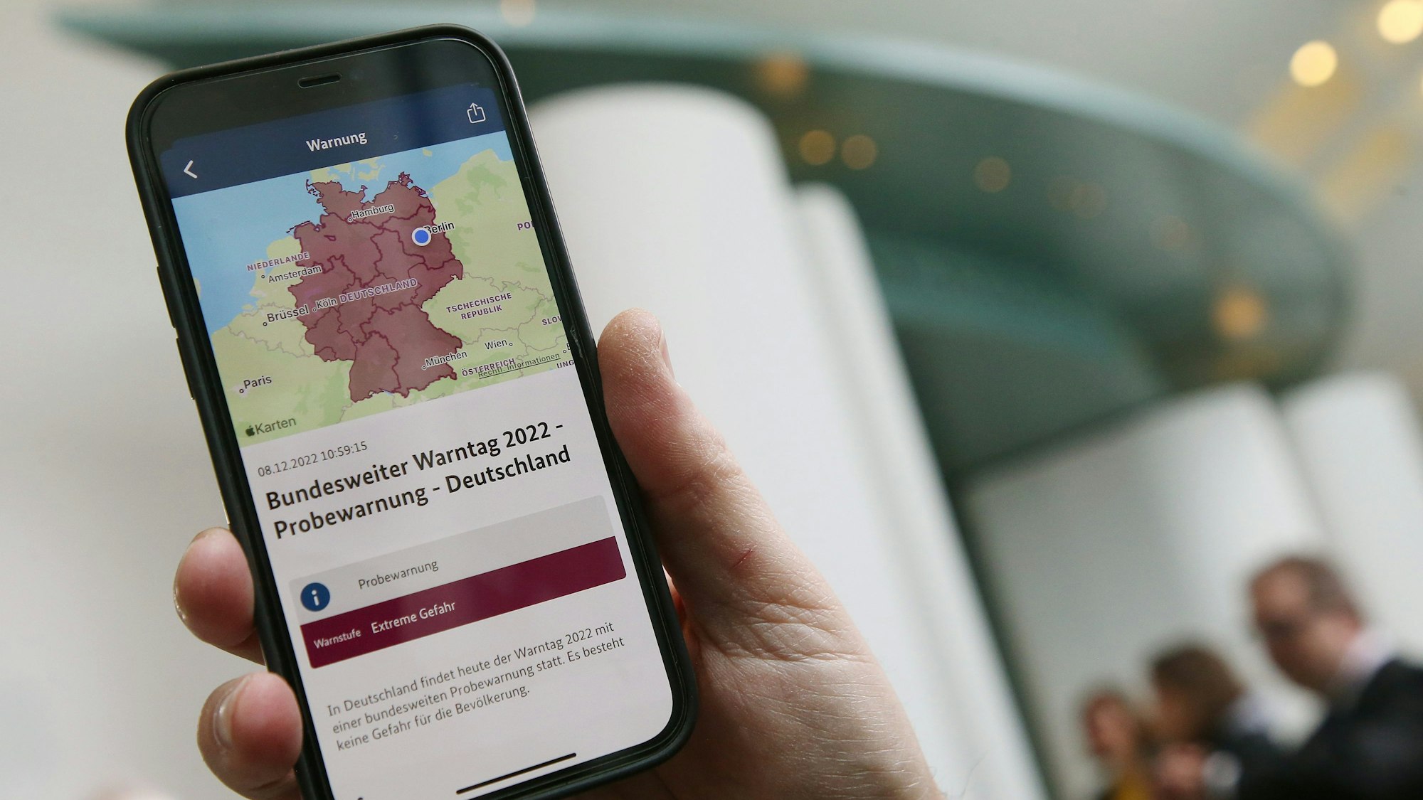 Ein Mitarbeiter des Bundeskanzleramts hält sein Mobiltelefon am Warntag 2022 im Foyer des Kanzleramtes in der Hand, wobei auf dem Display eine Probe-Warnmeldung zu sehen ist.