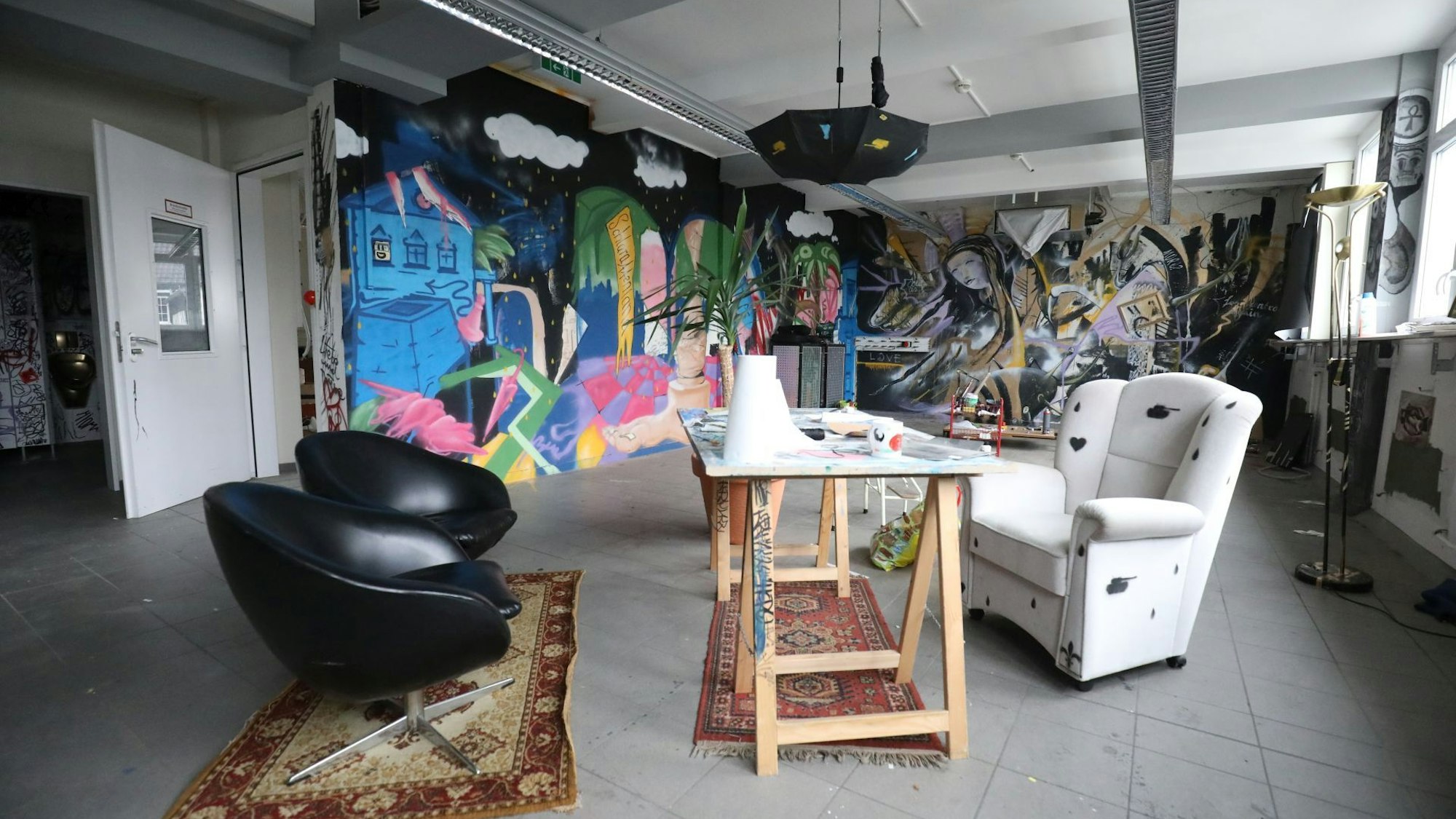 In einem Raum sind im Hintergrund die Wände mit großen Streetart-Bildern bemalt. Von der Decke hängt ein Regenschirm. Im Raum stehen zwei schwarze und ein weißer Sessel an einem Tisch aus Holzböcken.