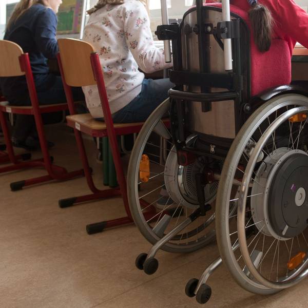 Eine Schülerin sitzt in einem Rollstuhl in einem Klassenraum. Sieht sie von hinten an einem grünen Tisch.