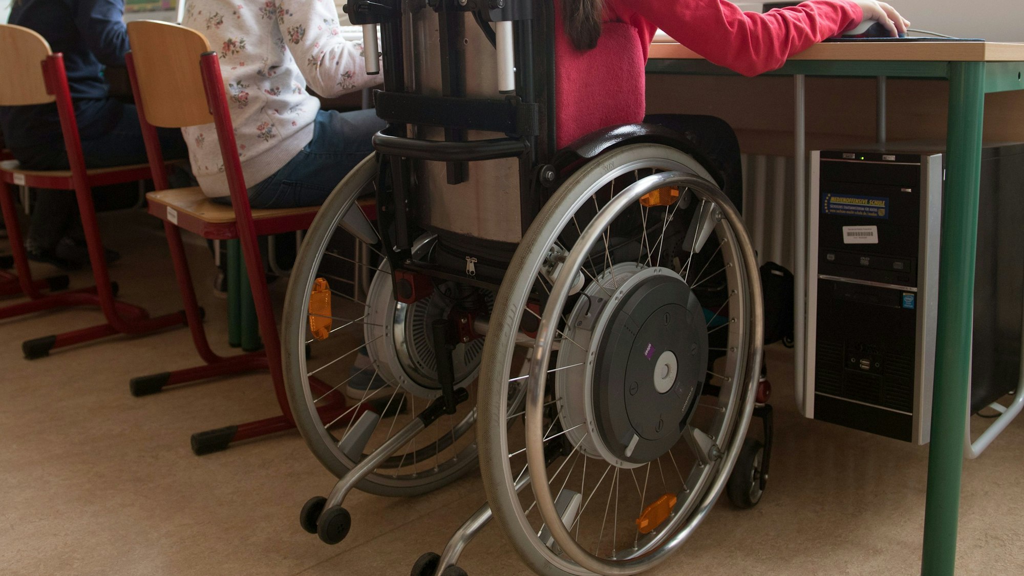Eine Schülerin sitzt in einem Rollstuhl in einem Klassenraum. Sieht sie von hinten an einem grünen Tisch.
