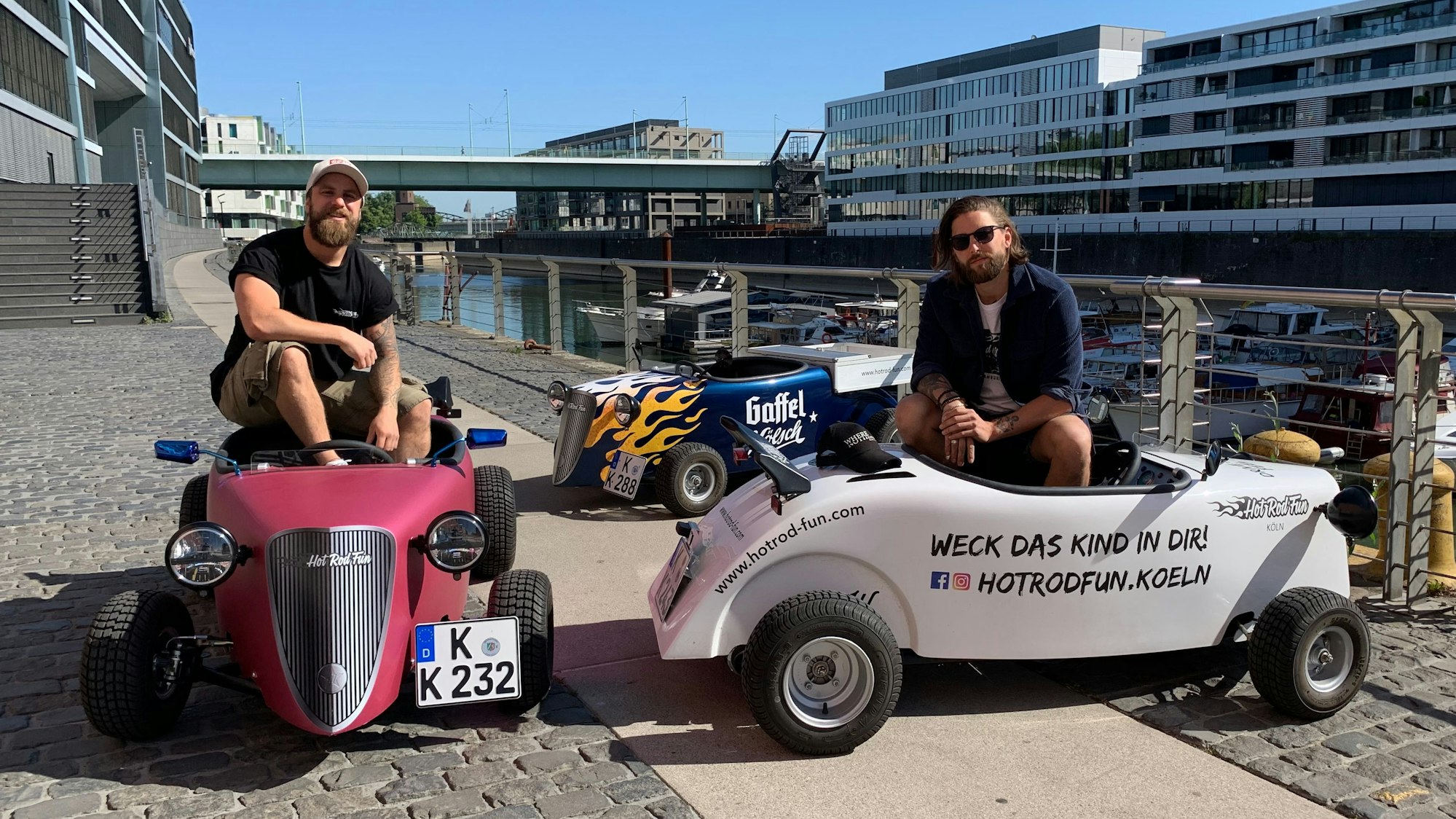 Zwei junge Unternehmer sitzen in den Miniatur-Nachbauten von Oldtimern im Kölner Rheinauhafen.