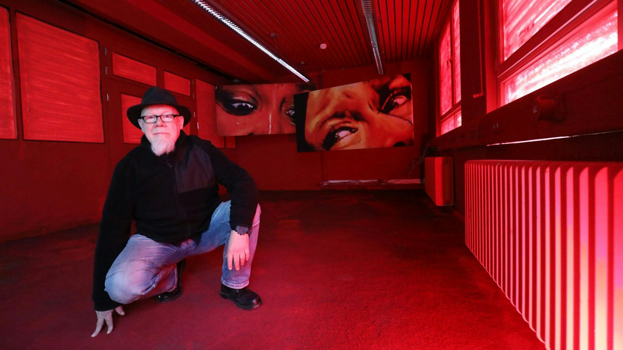 Ein Mann mit Hut kniet in einem komplett roten Raum, im Hintergrund hängen zwei großformatige Fotos, die verängstige Frauenaugen zeigen.