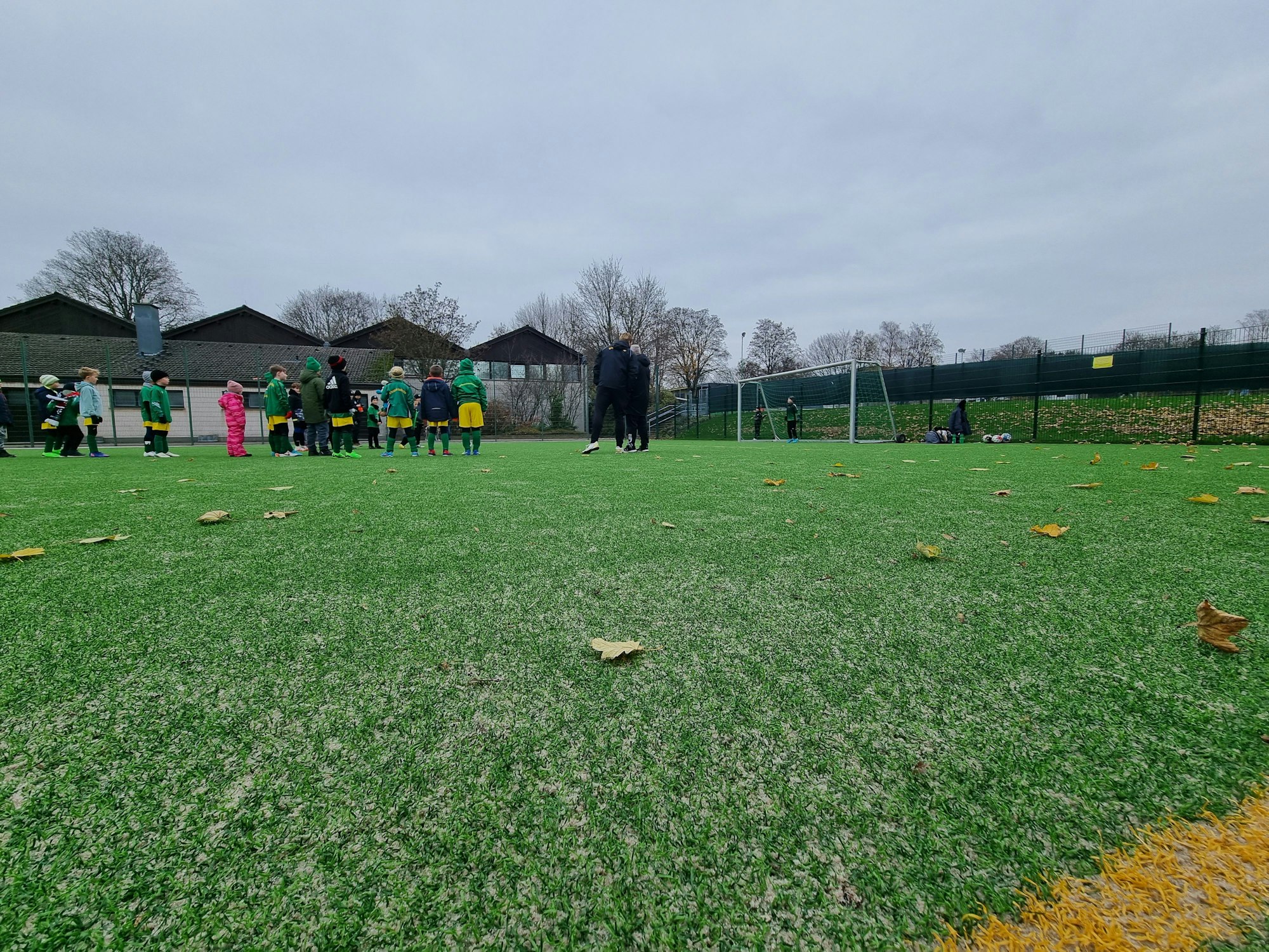 Nahaufnahme des grünen Kunstrasens, Teile der gelben Seitenlinie sind zu sehen. Im Hintergrund stehen Trainer und Kinder.