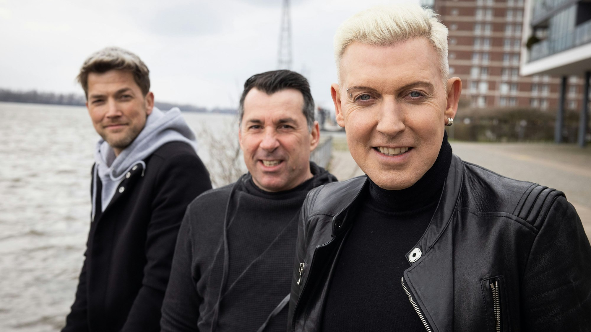 Drei Männer stehen am Rand eines Gewässers und blicken in die Kamera.