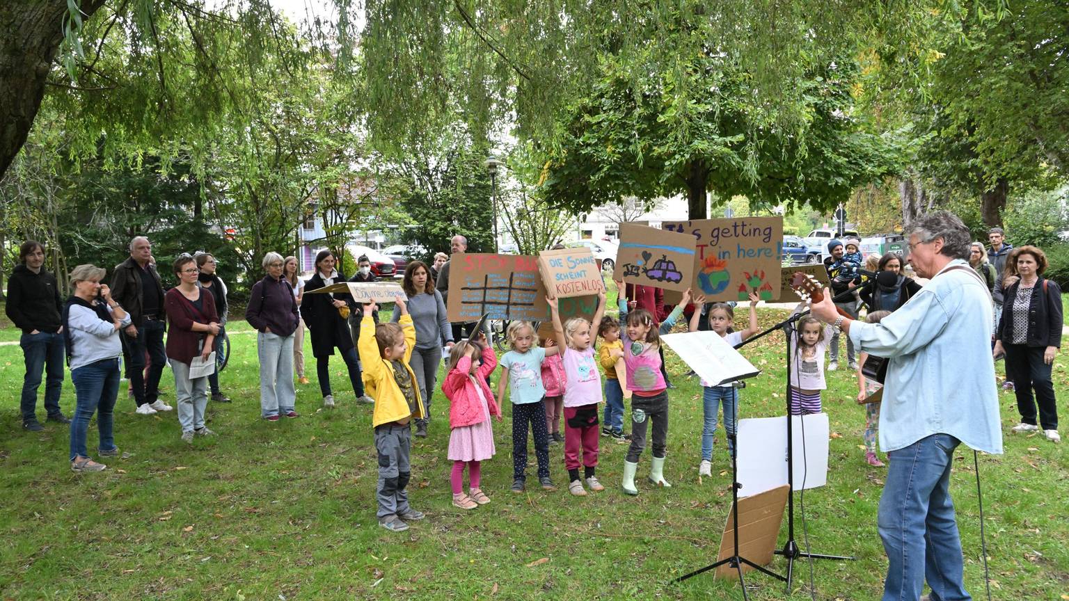 Auf einer Fridays for Future Demo in Rösrath halten Kinder Plakate hoch und ein Mann spielt Gitarre. Eltern stehen im Hintergrund.