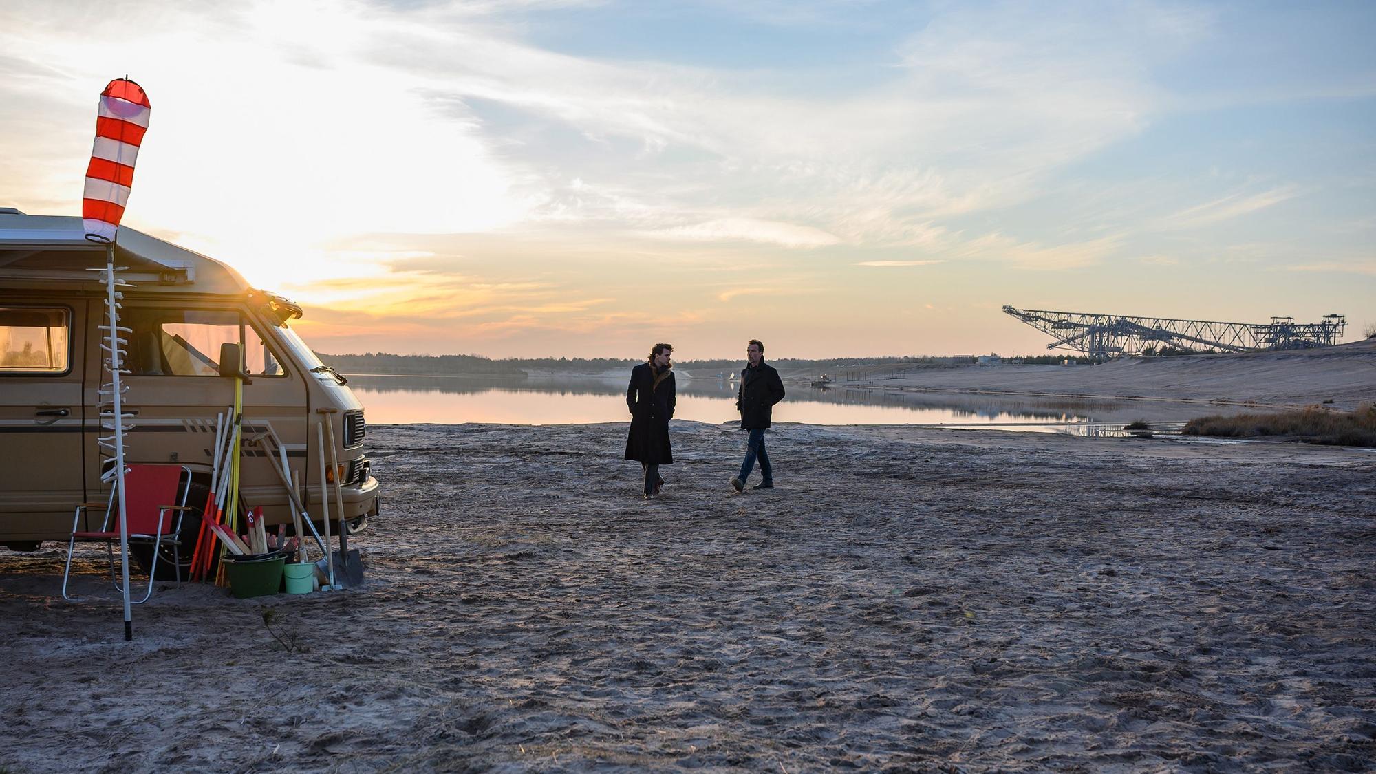 Ein weiter Ausblicam Strand eines Sees, im Hintergrund der ehemalige Tagebau. Links der Camper der Getöteten, in der Mitte das Ermittlerduo.