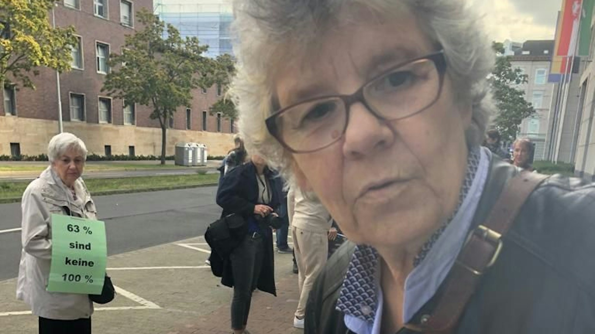 Renate Okrent guckt in die Kamera, sie trägt eine blaue Bluse, schwarze Jacke und eine Umhängetasche. Im Hintergrund stehen weitere Demonstrantinnen.