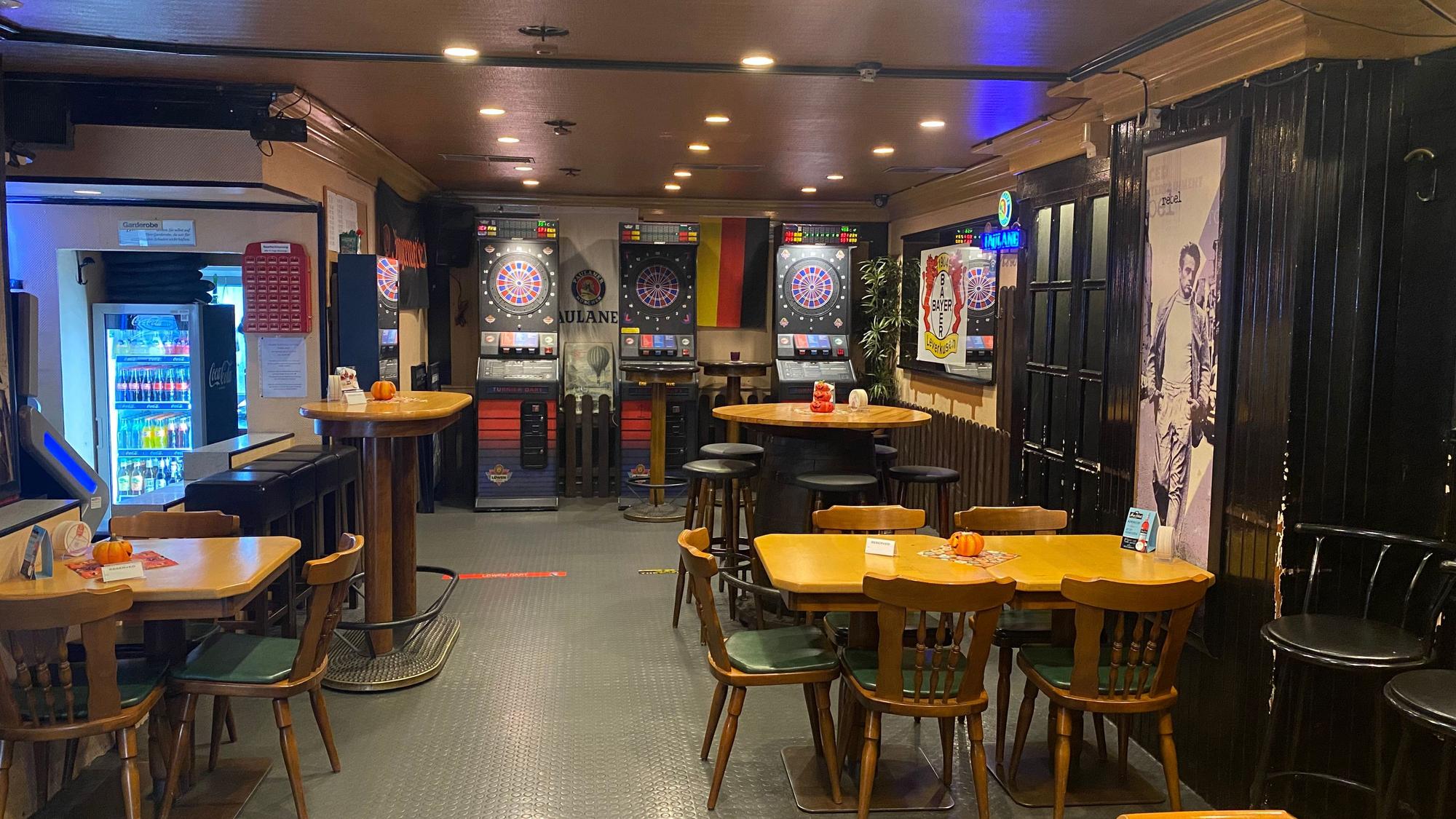 Blick in den leeren Schankraum der Kneipe Bierhaus. An einer Wand stehen drei Dartautomaten, an einer anderen hängt eine Fahne von Bayer 04 Leverkusen.