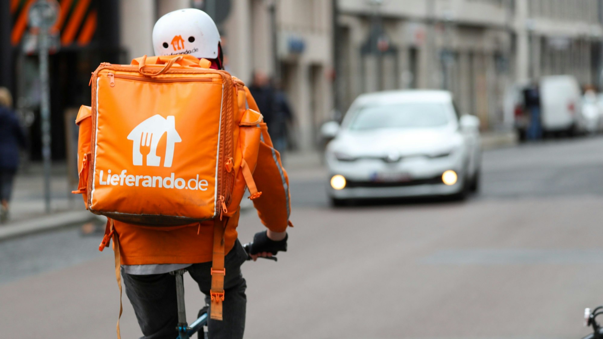 Ein Fahrradbote vom Lieferdienst „Lieferando“ fährt durch die Innenstadt. Auf dem Rücken trägt er einen orangefarbenen Rucksack.