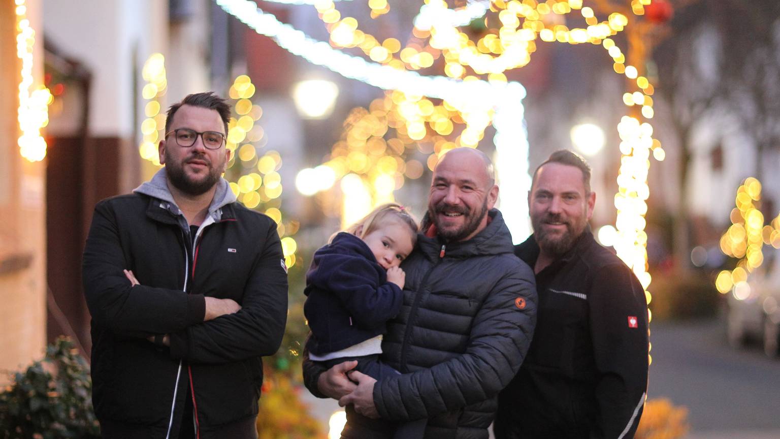 Die Initiatoren der Weihnachtsstraße (von links) Patrick Schübl, Daniel Thimm (mit Töchterchen Flora) und Thorsten Simmerkus stehen in der Dämmerung vor dem Lichterglanz an Bäumen und Vorgärte.