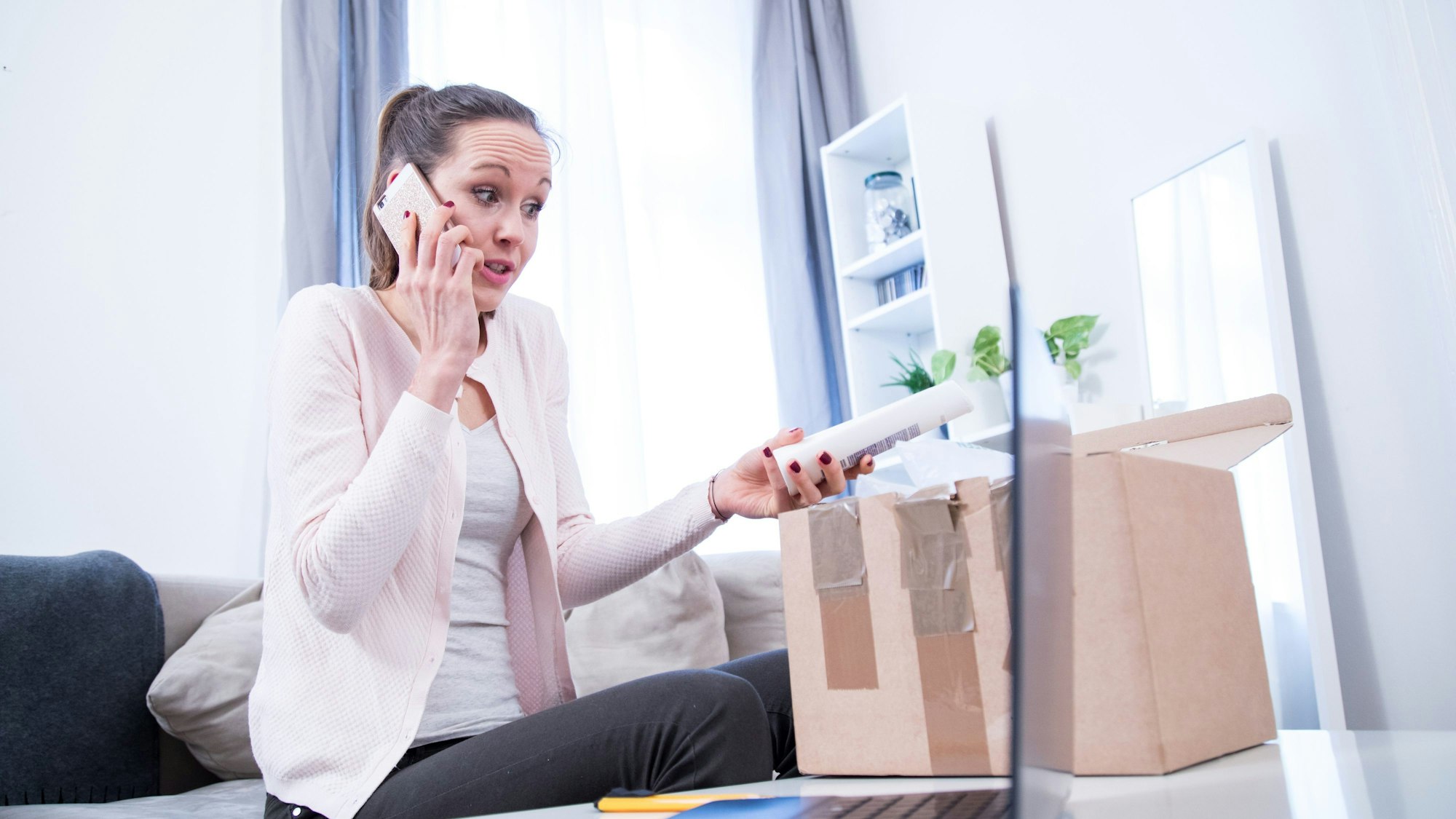 Eine Frau sitzt neben einem Paket, hält eine Verpackung in der Hand und telefoniert währenddessen.