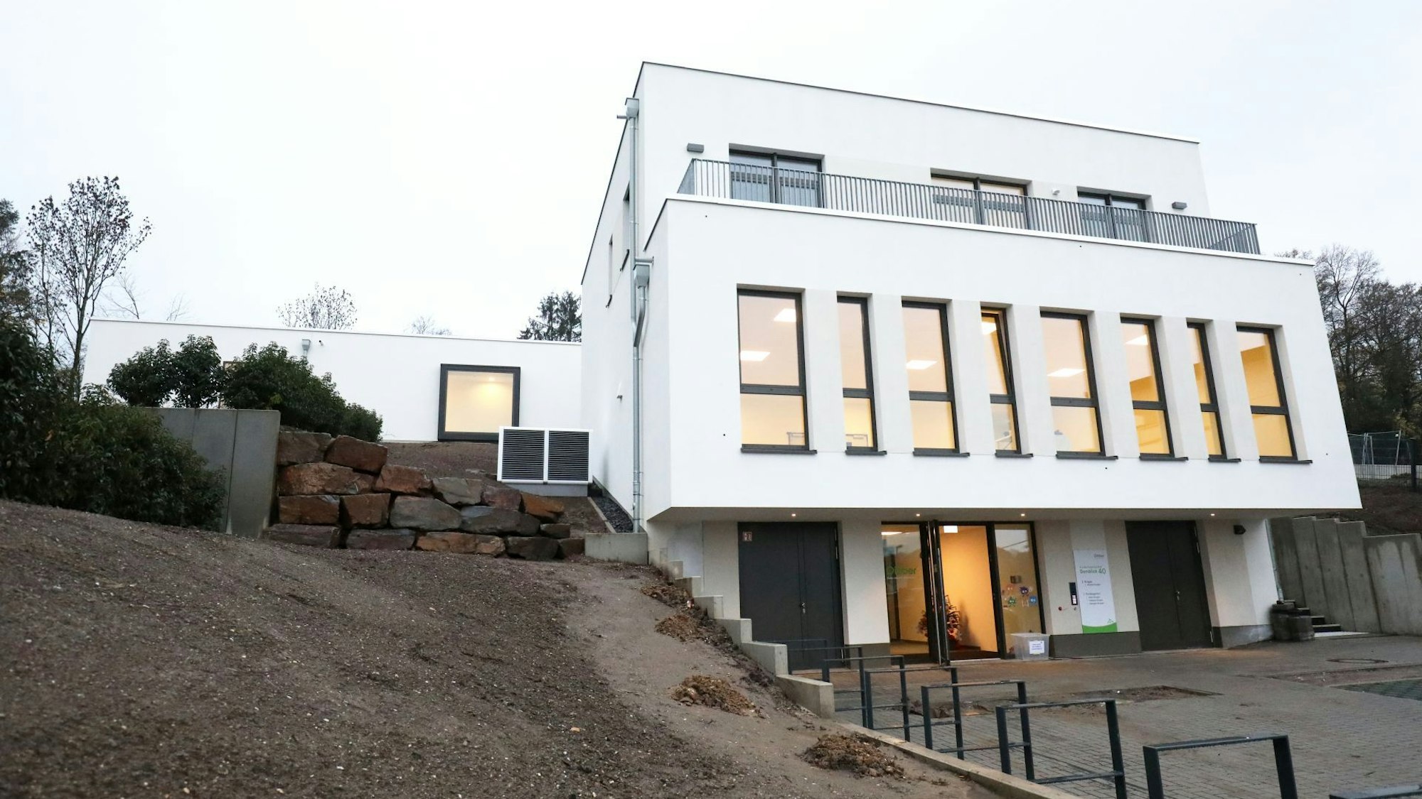 Das weiße KiTa-Gebäude mit hohen Fenstern ist hell erleuchtet.