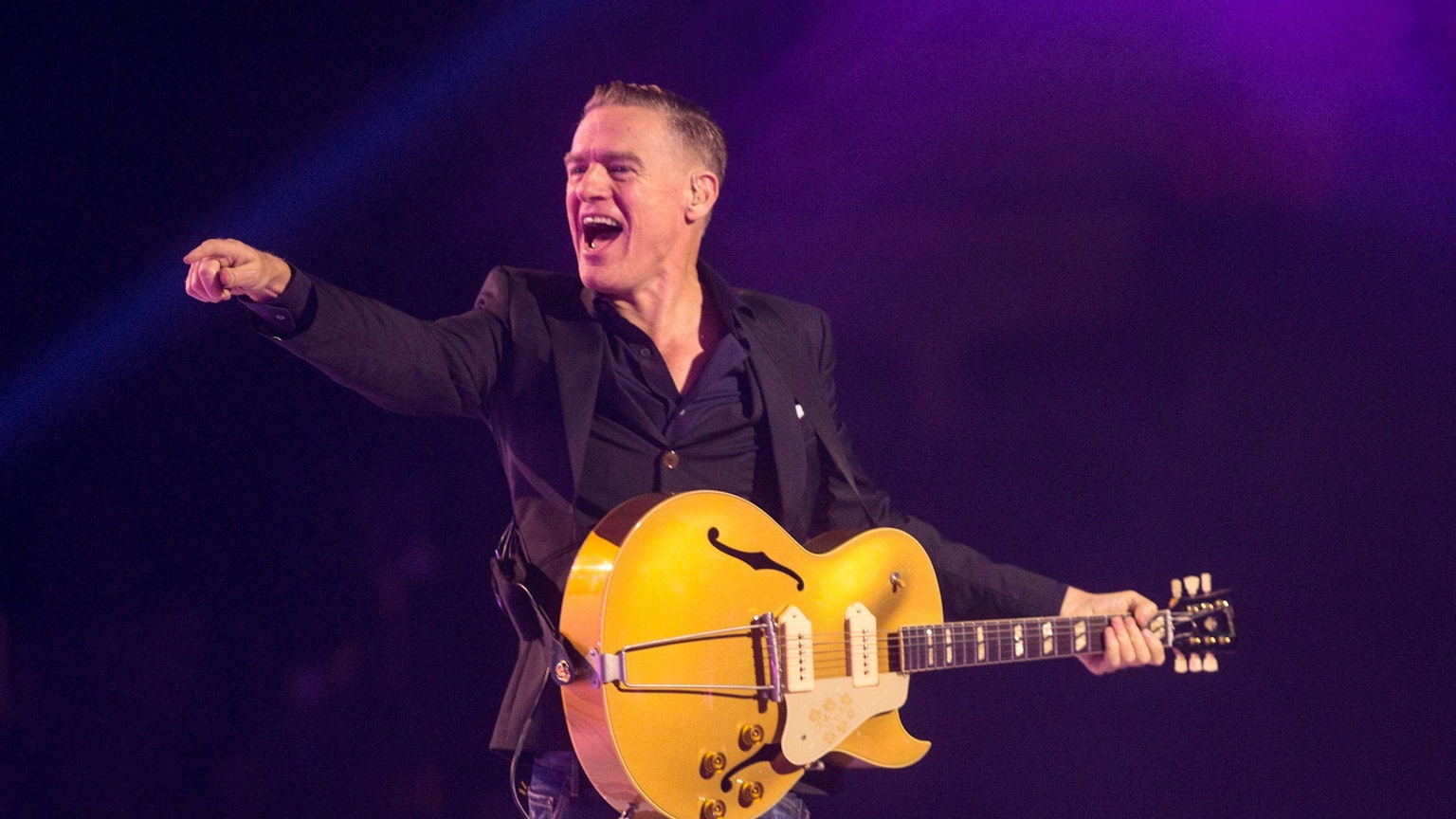 Bryan Adams bei einem Konzert mit Gitarre. Er streckt den Finger Richtung Publikum und lacht.
