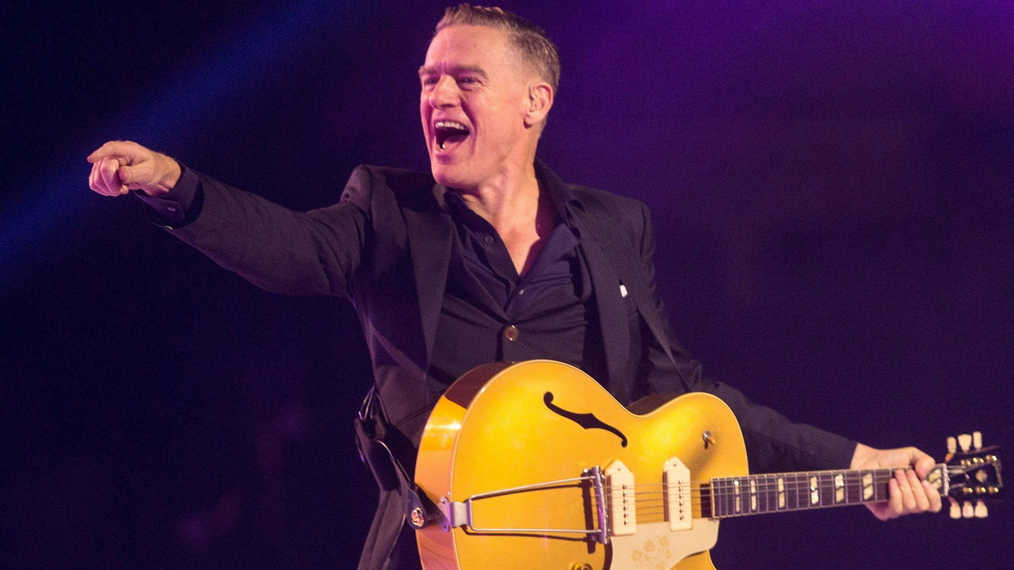 Bryan Adams bei einem Konzert mit Gitarre. Er streckt den Finger Richtung Publikum und lacht.
