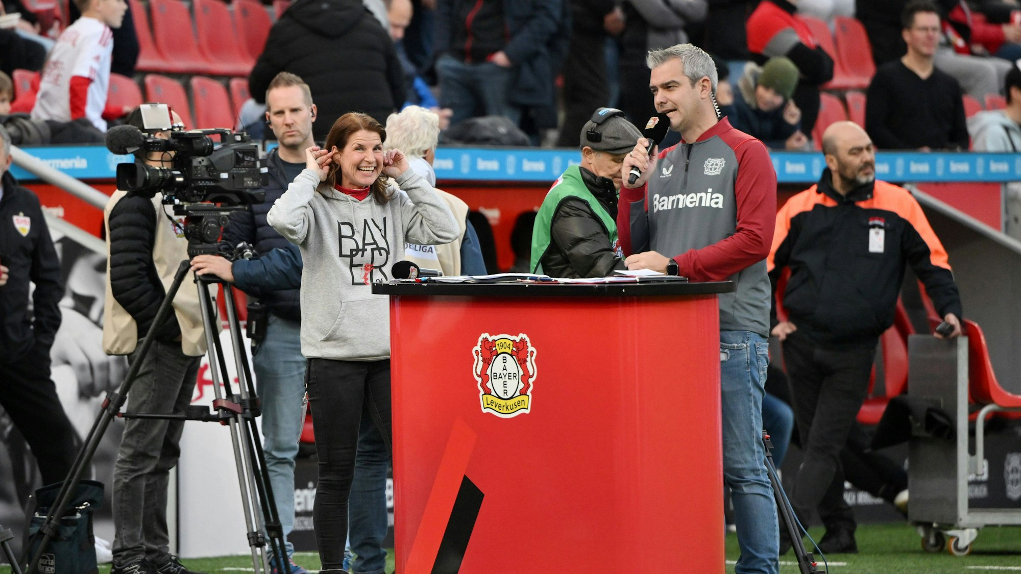 Stadionsprecher Tobias Ufer steht mit seiner Kollegin, Stadionsprecherin Petra Dahl, an einem roten Pult auf dem Spielfeld und spricht in ein Mikrofon.