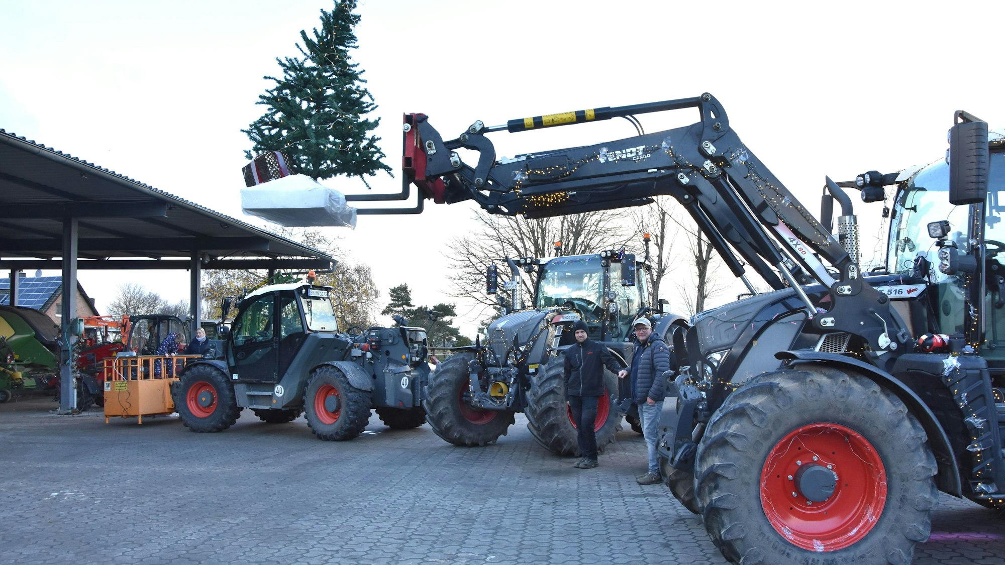 Mehrere Traktoren stehen auf einem Hof, zwei Männer in der Mitte. Ein Traktor hebt einen Tannenbaum.