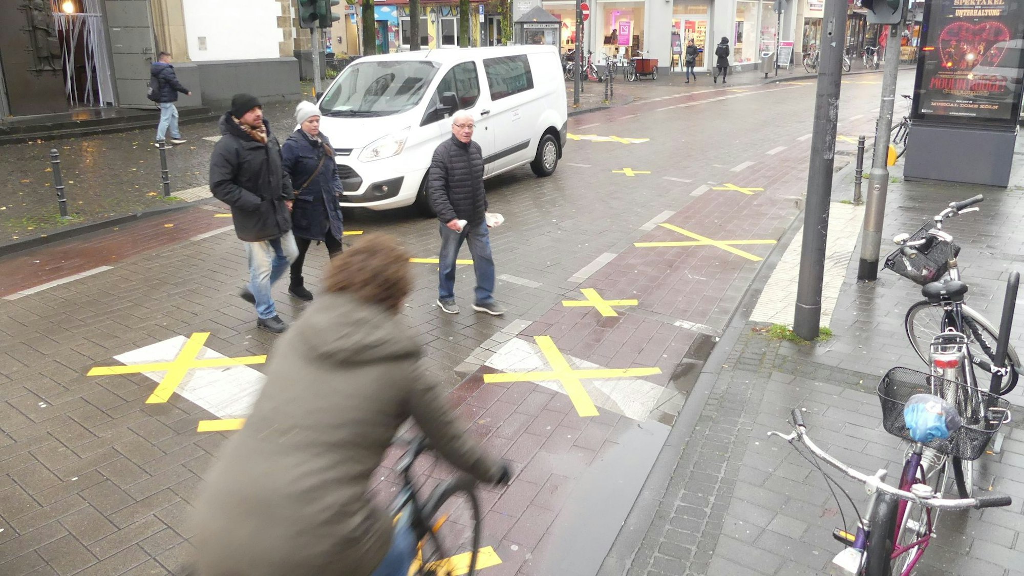 Auf der Venloer Straße sind Radwegmarkierungen und Haltemarkierungen für Autos vor Fußgängerüberwegen mit gelben Klebestreifen überklebt. Was sie bedeuten, ist unklar. Ein Fahrradfahrer nutzt den überklebten Radweg, Fußgänger die überklebte Straßenquerung.