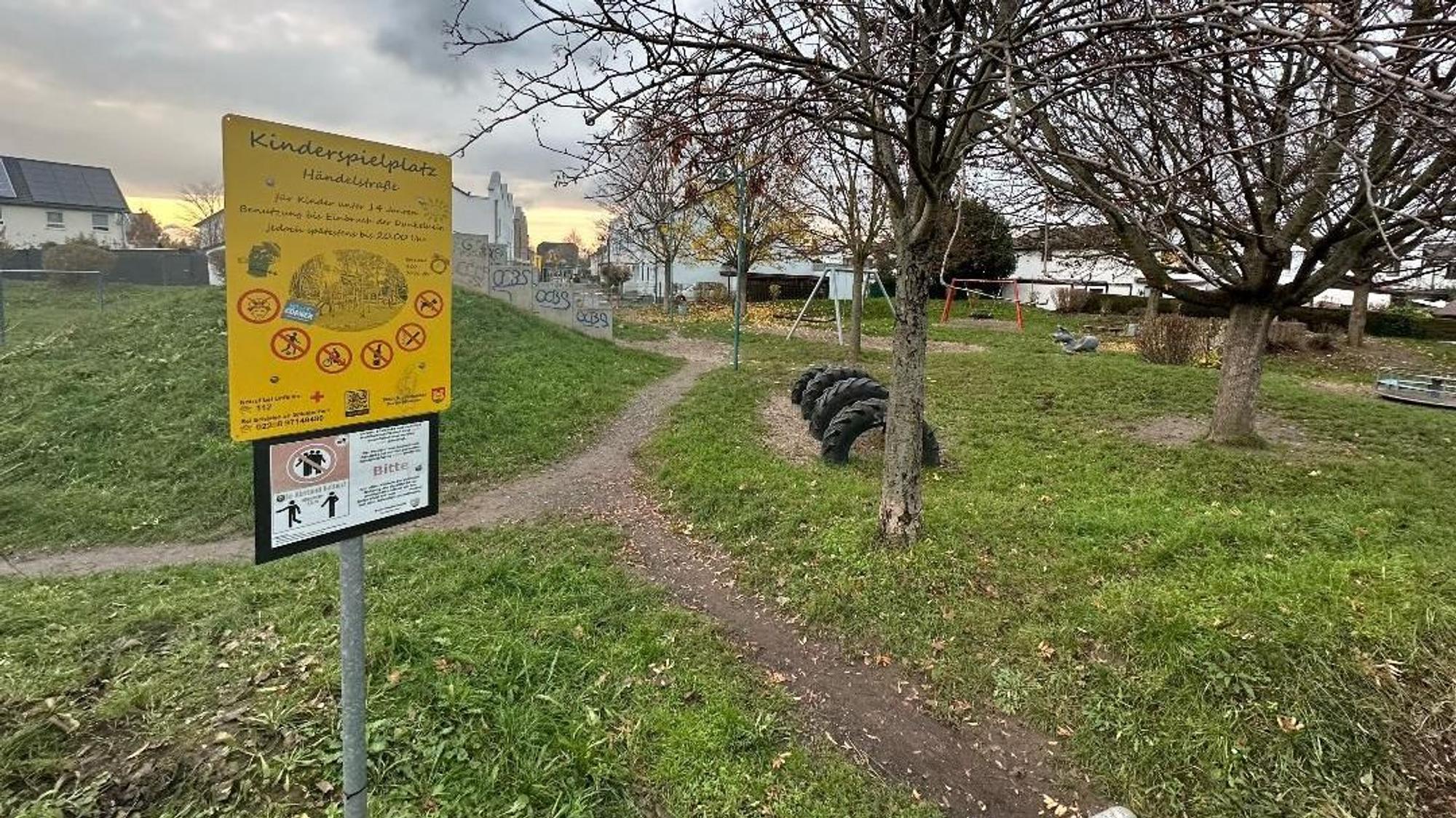 Ein leerer, heruntergekommener Spielplatz in Niederkassel. Im Vordergrund ist ein gelbes Schild, auf dem "Kinderspielplatz Händelstraße" steht. Im Hintergrund sind Spielgeräte wie Schaukeln, Reifen und Wippen zu sehen.
