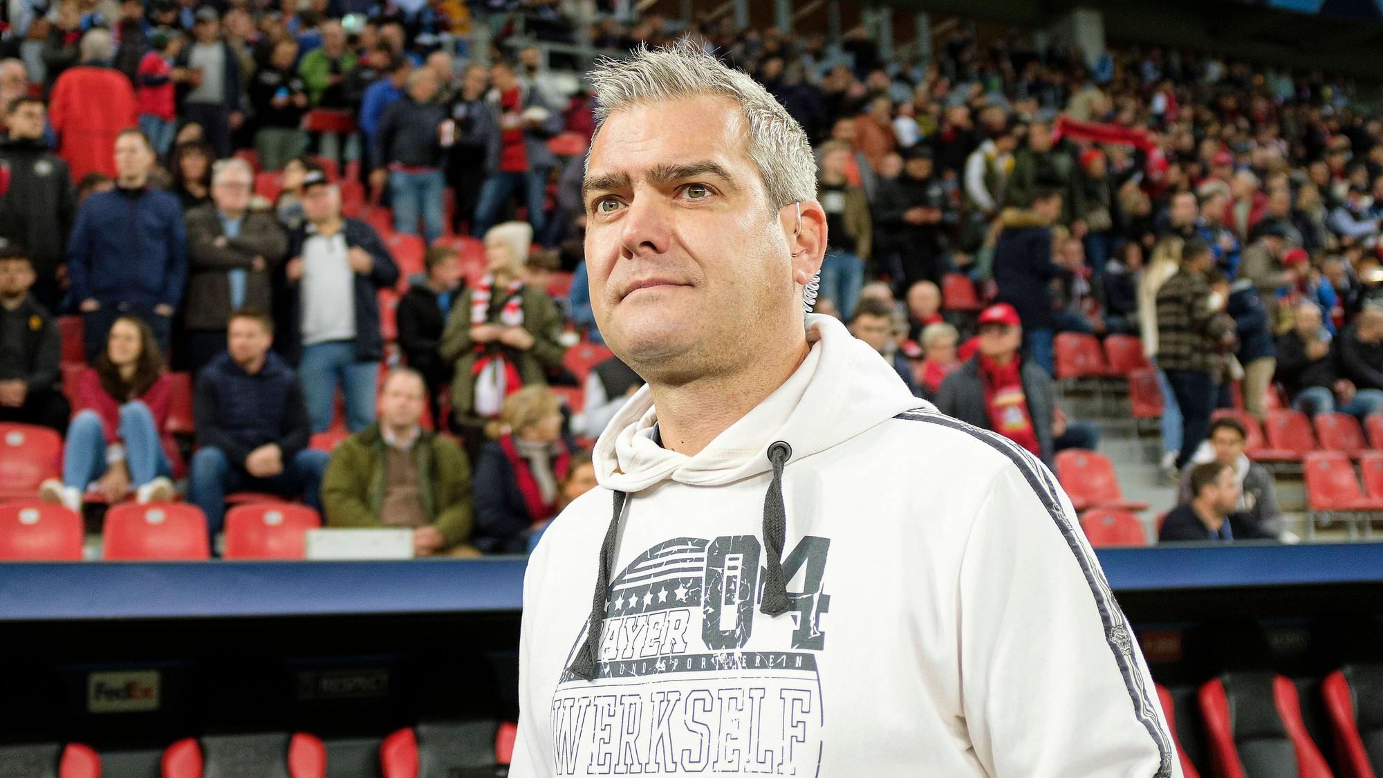 Stadionsprecher Tobias Ufer steht auf dem Spielfeld der Bayarena vor einer Tribüne, auf der Zuschauer sitzen.