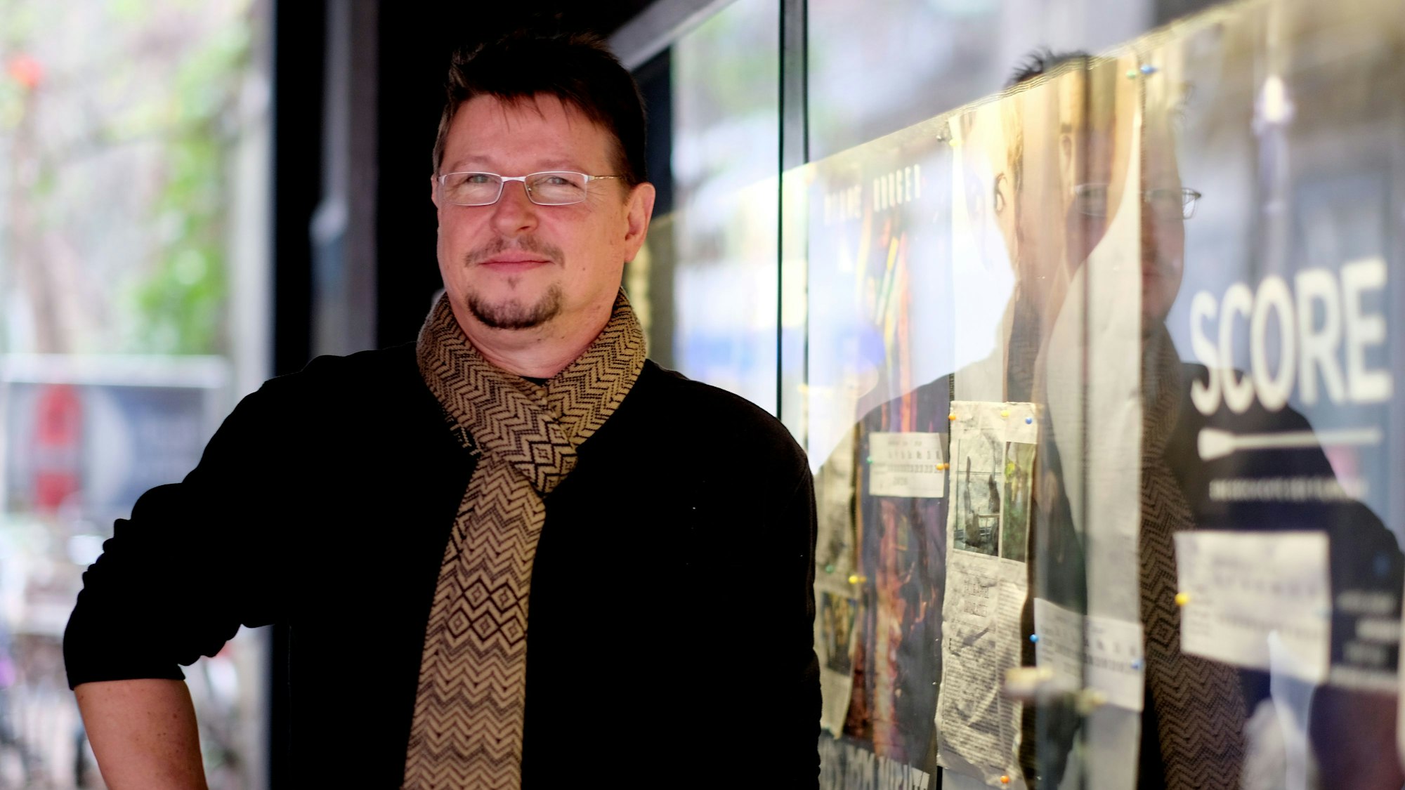 Christian Schmalz steht mit schwarzem Pullover und gemustertem Schal vor Filmplakaten an seinem Kölner Kino Off Broadway an der Zülpicher Straße.
