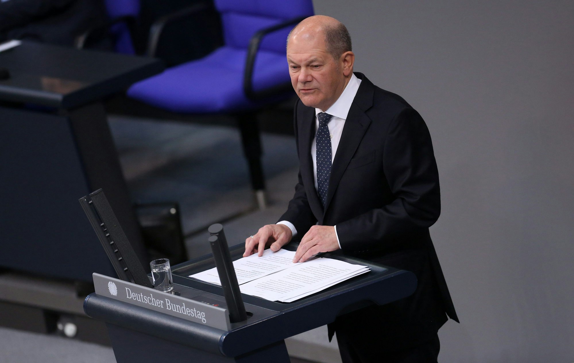 Bundeskanzler Olaf Scholz spricht im Deutschen Bundestag. Vor ihm steht ein Glas Wasser, er hat seine Rede ausgedruckt vor sich liegen.