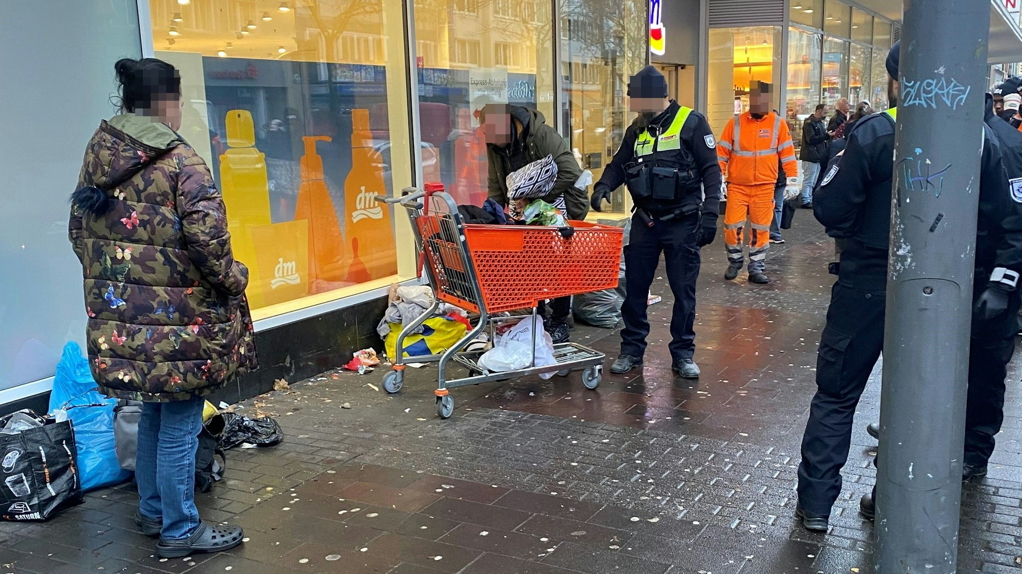 Ein Mitarbeiter des Ordnungsamts steht neben einem Obdachlosen, der vor einem Einkaufswagen steht.