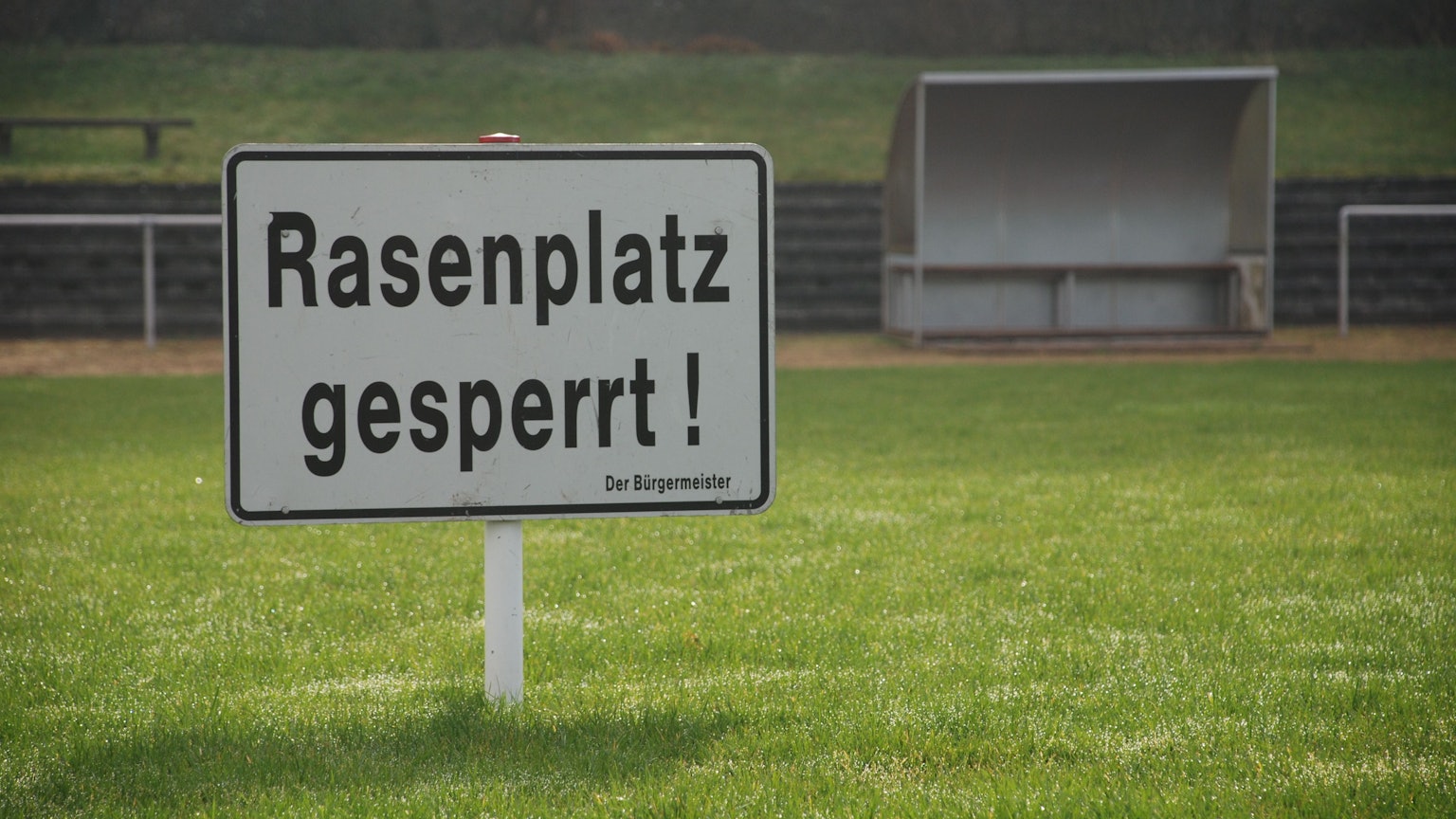 Auf einem Rasenplatz steht das Schild „Rasenplatz gesperrt“. Im Hintergrund ist eine Bank mit Überdach zu sehen.