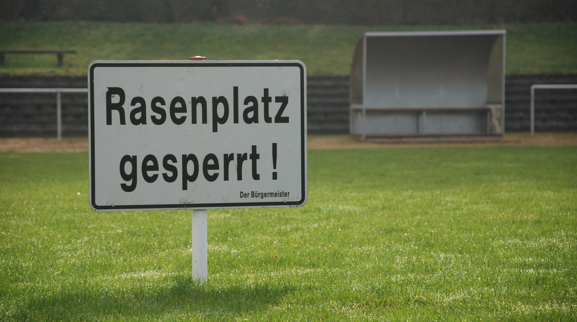 Auf einem Rasenplatz steht das Schild „Rasenplatz gesperrt“. Im Hintergrund ist eine Bank mit Überdach zu sehen.