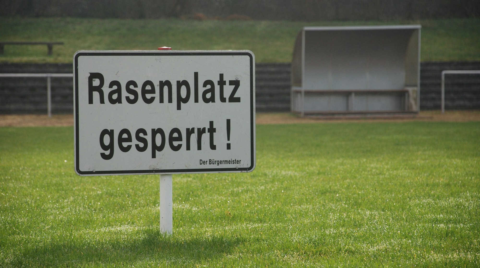 Auf einem Rasenplatz steht das Schild „Rasenplatz gesperrt“. Im Hintergrund ist eine Bank mit Überdach zu sehen.