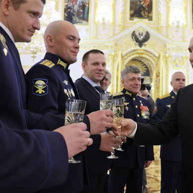 Der russische Staatspräsident Wladimir Putin beglückwünscht mehrere Personen während einer Zeremonie am Vorabend des Tages der Helden des Vaterlandes im Kreml. Er trägt einen Anzug und hat ein Champagner-Glas in der Hand.