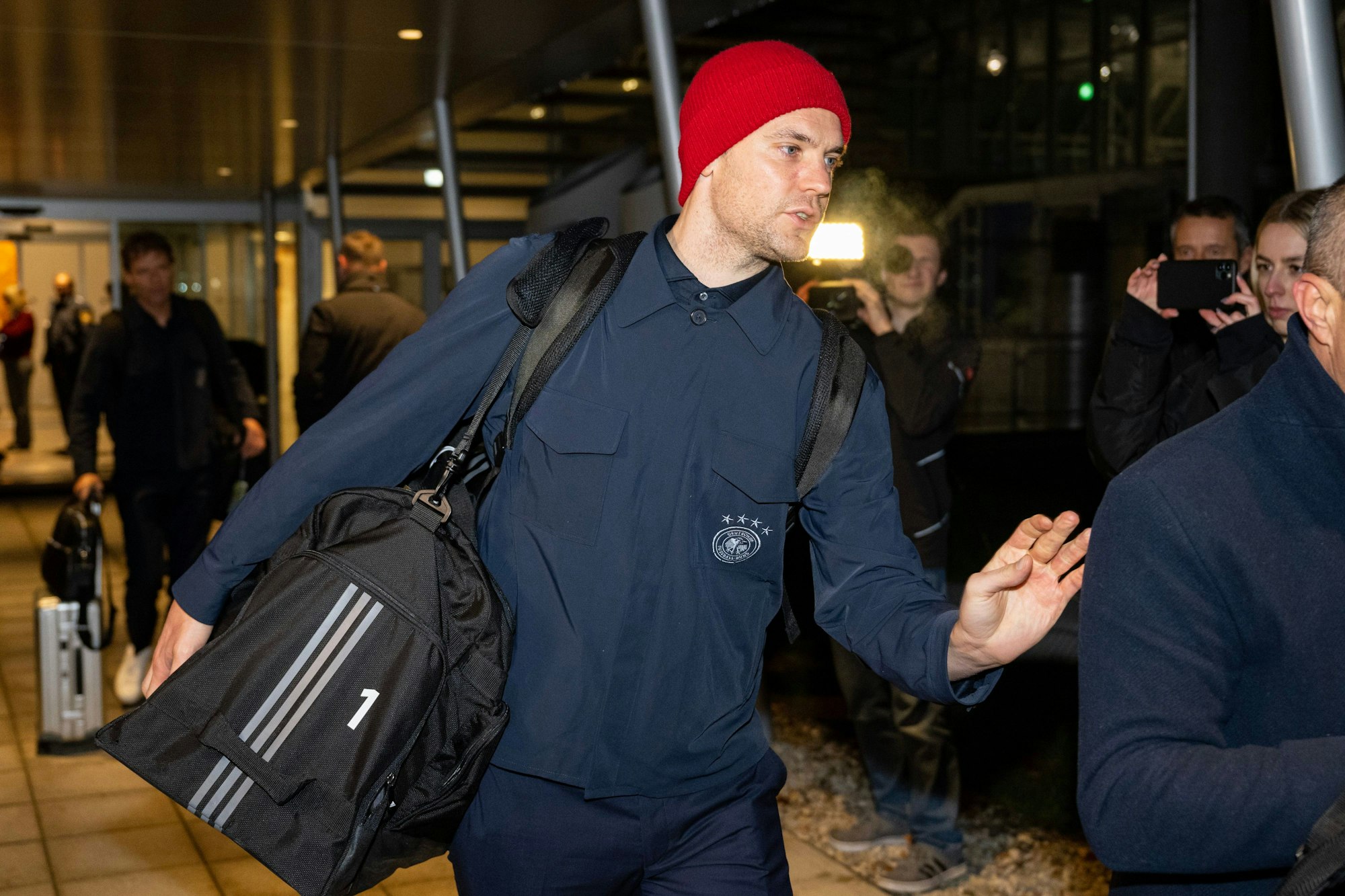 Manuel Neuer in blauem Trainingsanzug und mit roter Mütze am Flughafen in München.